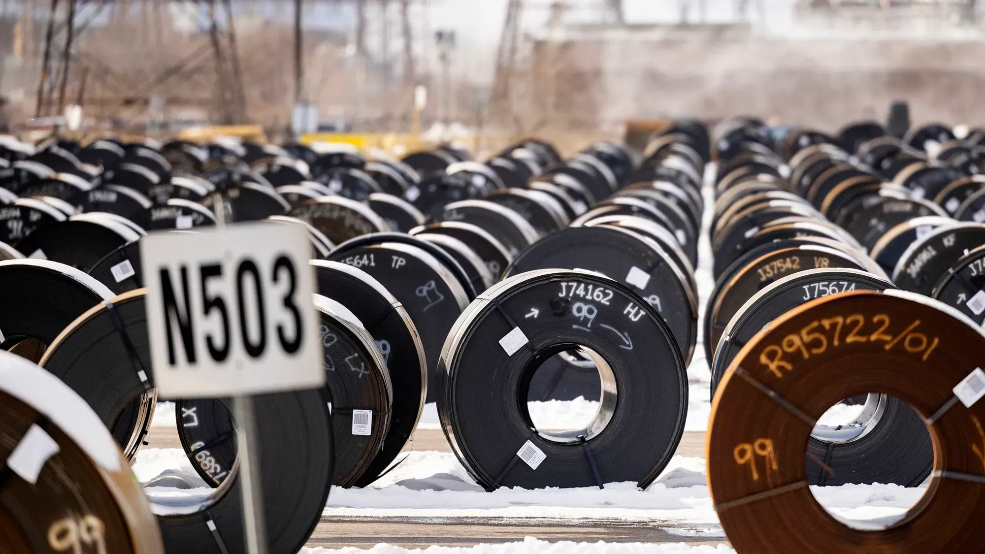 صفوف من لفائف الصلب في موقع تخزين، مع علامة تشير إلى الموقع، تعكس تأثير الرسوم الجمركية على الصناعة الأمريكية.