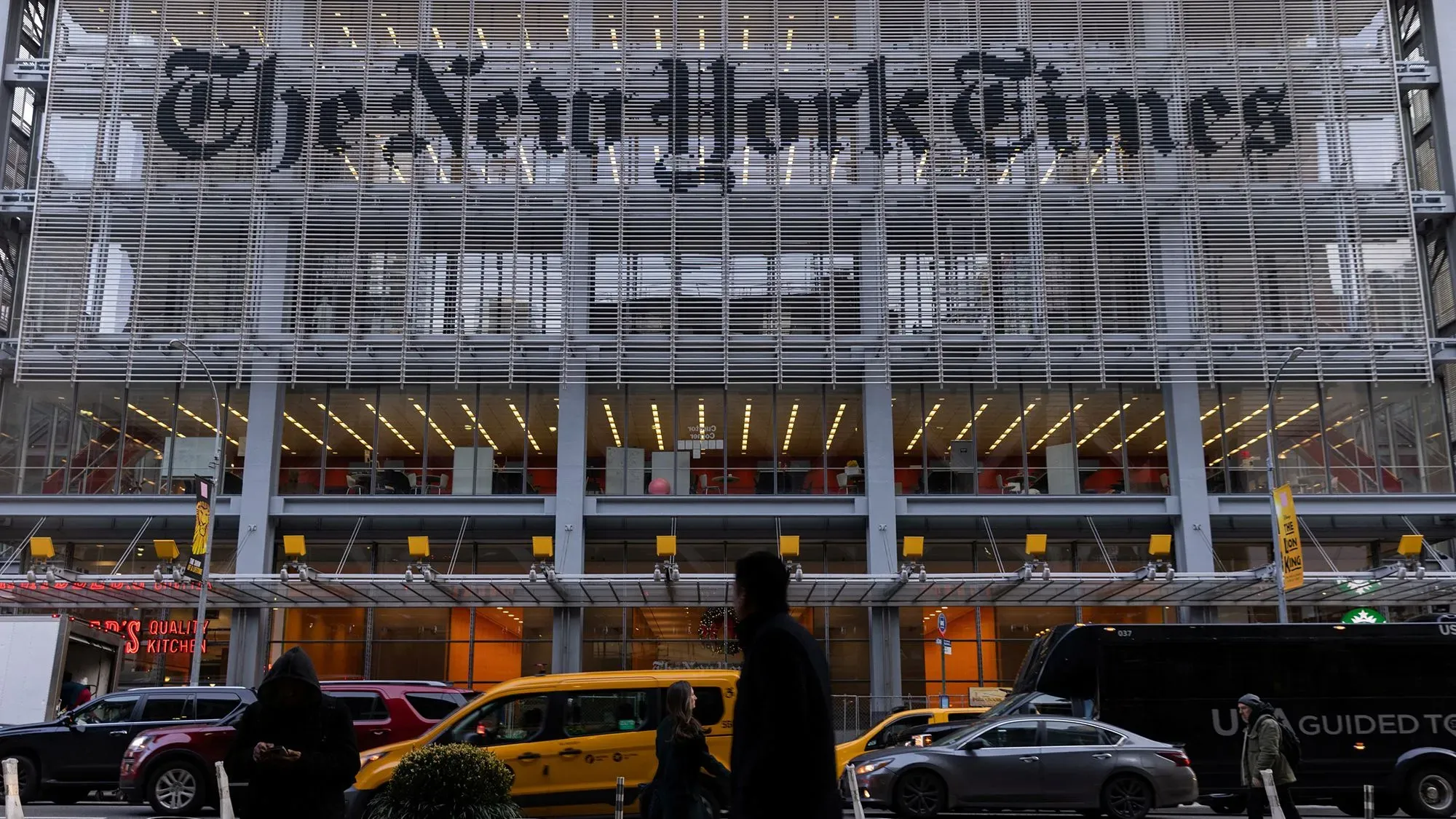 مبنى صحيفة نيويورك تايمز في نيويورك، مع لافتة كبيرة تحمل اسم الصحيفة، بينما يمر الناس والسيارات في الشارع.