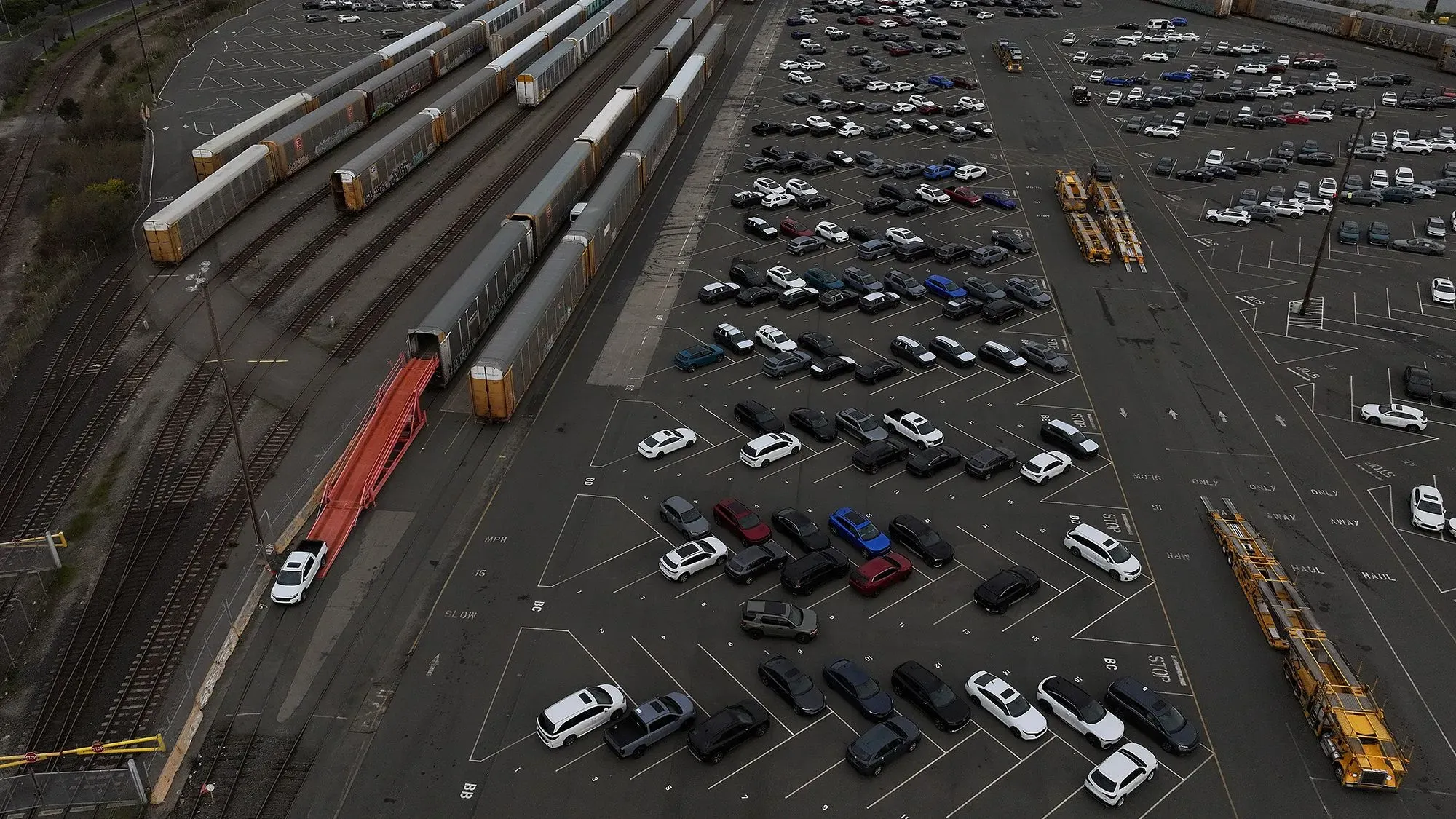 صورة جوية لموقف سيارات واسع يحتوي على العديد من السيارات، مع وجود قطارات شحن في الخلفية، تعكس تأثير الرسوم الجمركية على صناعة السيارات.