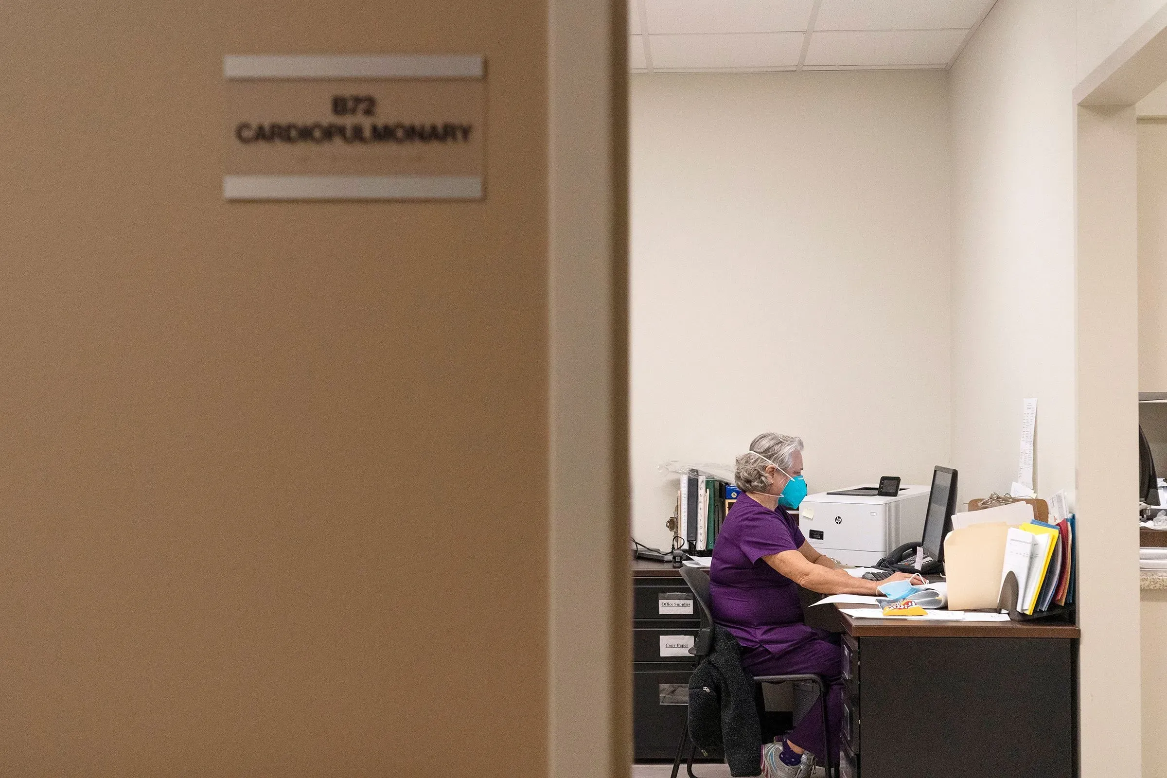 امرأة ترتدي قناعًا جراحيًا تجلس أمام جهاز كمبيوتر في مكتب طبي، مع علامة تشير إلى قسم القلب والرئة.