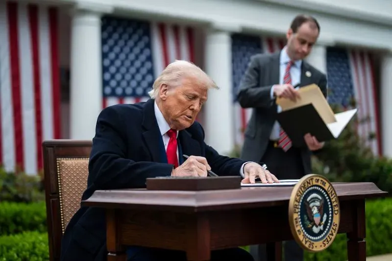 ترامب يوقع وثيقة في حديقة البيت الأبيض، مع وجود علم أمريكي خلفه، وسط أجواء تعكس التوترات الاقتصادية والسياسية.