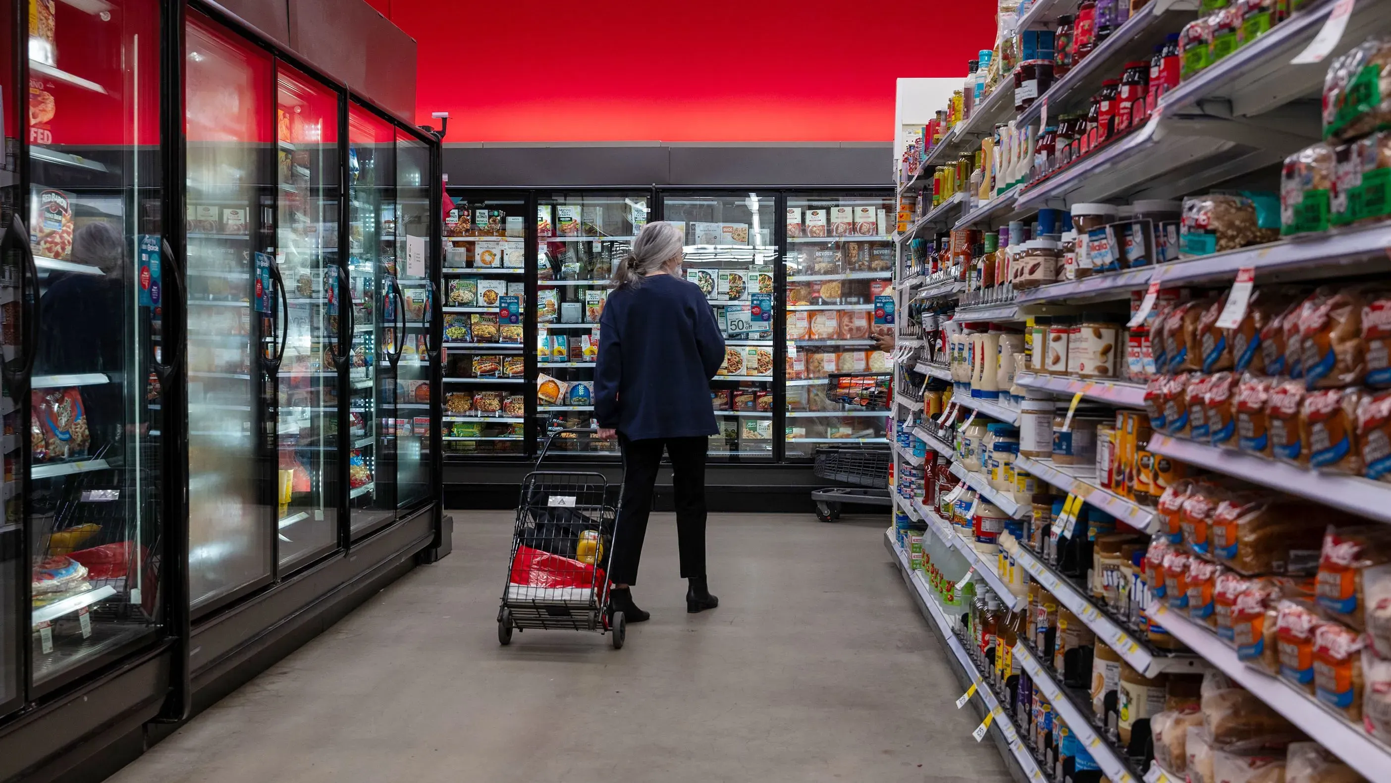 امرأة مسنّة تتسوق في متجر، تقف أمام قسم المجمدات، مع وجود أرفف مليئة بالمنتجات الغذائية على الجانبين، تعكس تأثير ارتفاع الأسعار على المستهلكين.