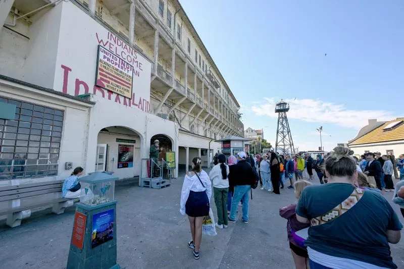 طابور طويل من الزوار أمام مبنى سجن الكاتراز التاريخي، حيث يظهر برج المراقبة في الخلفية، مع لافتات ترحيب بالزوار.