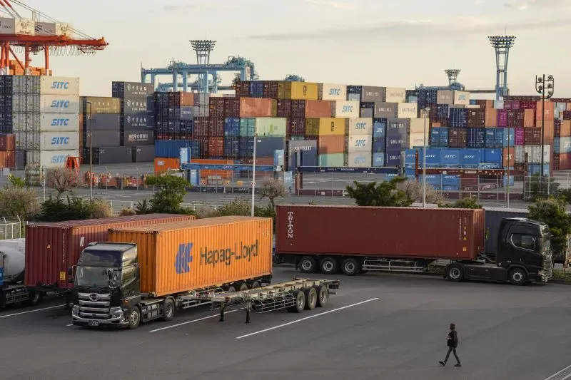 شاحنة تنقل حاويات شحن في ميناء ياباني، مع خلفية مملوءة بالحاويات الملونة، تعكس حركة التجارة الدولية.