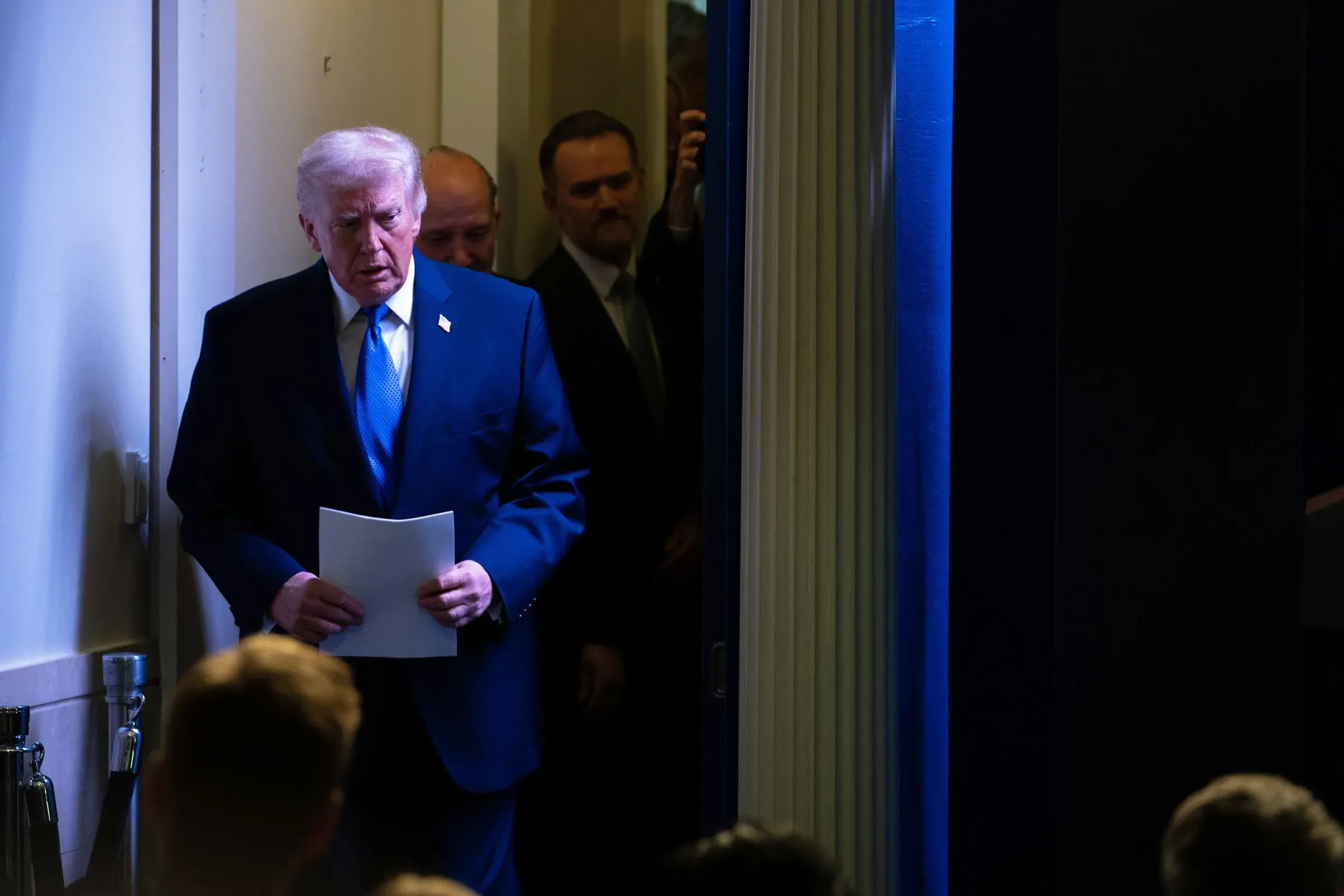 ترامب يدخل قاعة المؤتمر حاملاً ورقة، مع إضاءة خافتة خلفه، في سياق حديثه عن الرسوم الجمركية وتأثيرها على الاقتصاد الأمريكي.