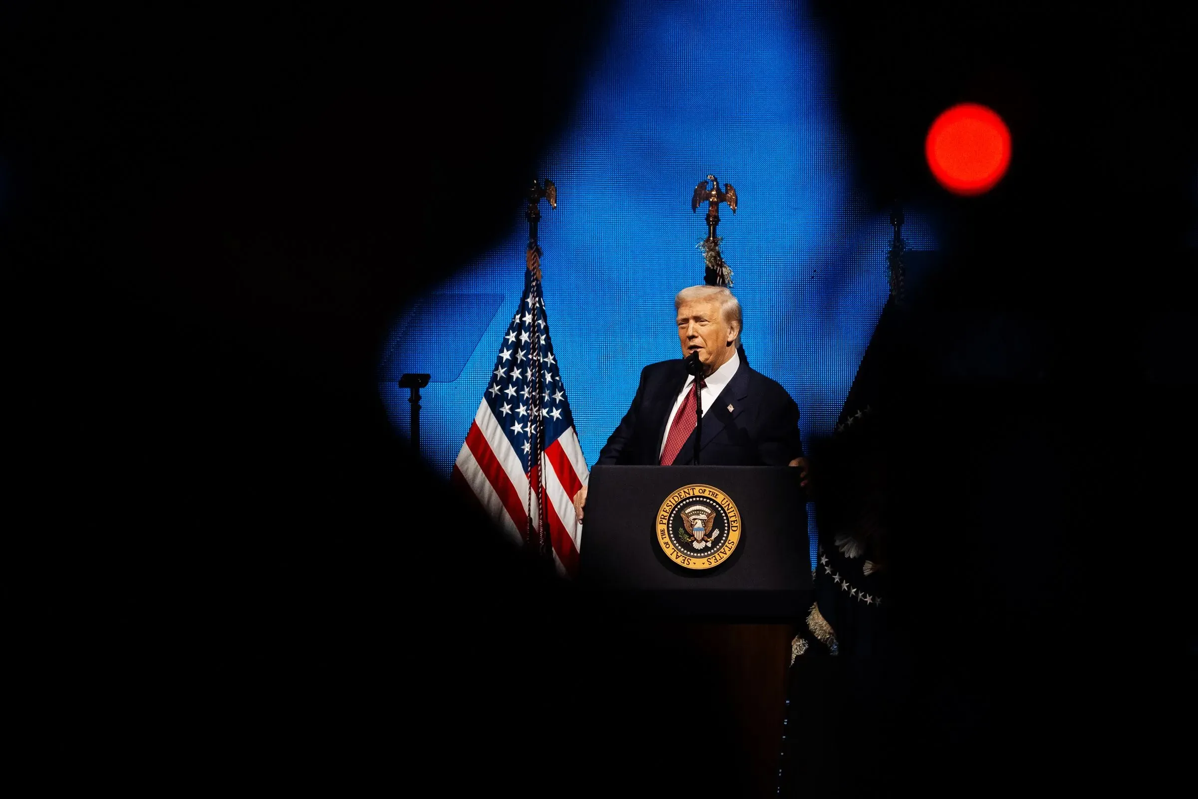 ترامب يتحدث في حدث سياسي، مع العلم الأمريكي خلفه، وسط أضواء خافتة، مما يعكس أجواء المنافسة الانتخابية المحتدمة.