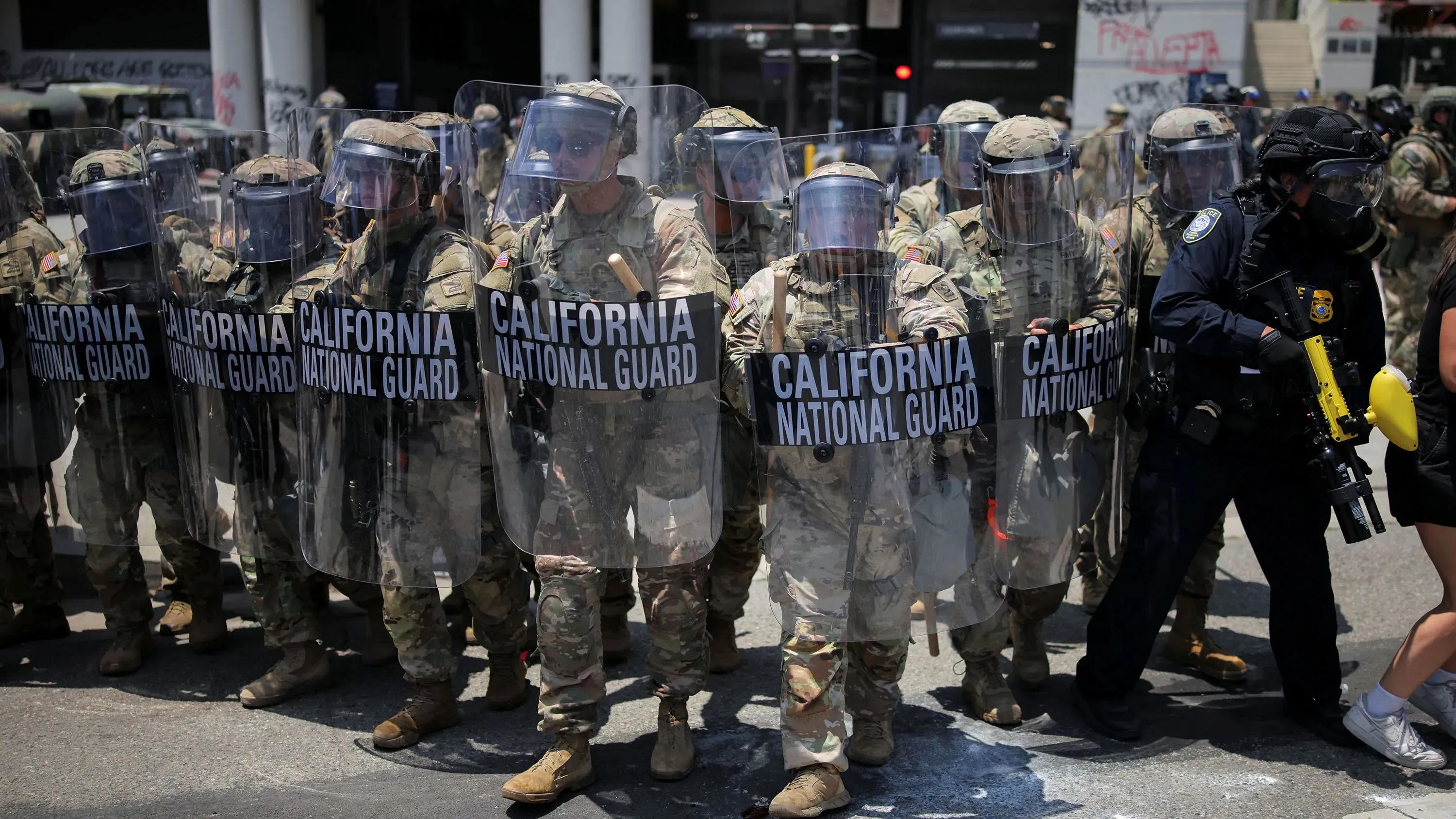 جنود من الحرس الوطني في كاليفورنيا يرتدون دروعًا واقية ويحملون لافتات، أثناء مواجهة الاحتجاجات التي تلت مداهمات إدارة الهجرة والجمارك.