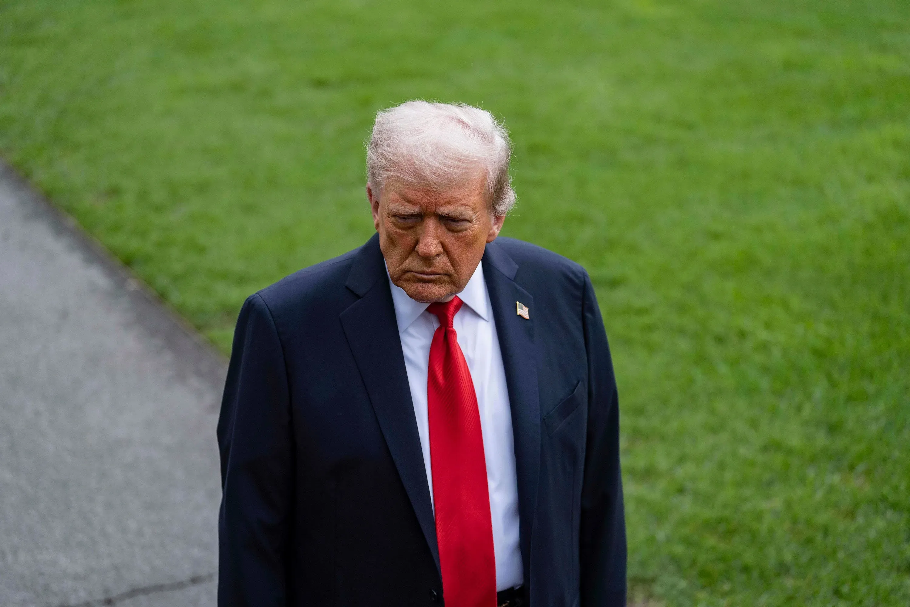 ترامب يسير بخطوات واثقة، مرتديًا بدلة داكنة وربطة عنق حمراء، مع تعبير وجه يعكس الجدية، في خلفية خضراء تعزز التركيز على ملامحه.
