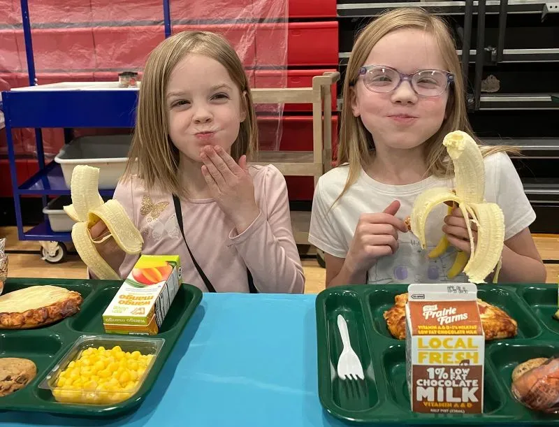 طفلتان تستمتعان بوجبة مدرسية، تتضمن الموز والحليب، تعكسان أهمية التغذية في بيئة التعليم.