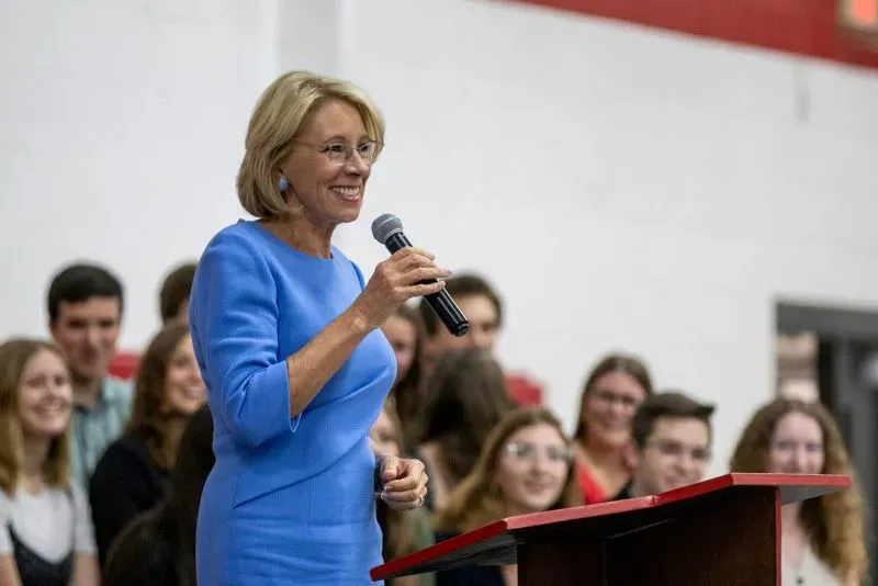 وزيرة التعليم السابقة بيتسي ديفوس تتحدث في حدث مدرسي، محاطة بجمهور من الطلاب، في إطار مناقشة حول برنامج القسائم المدرسية.