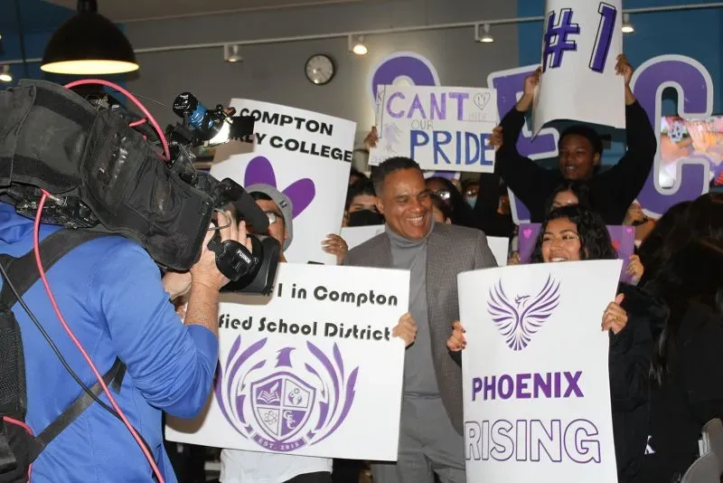 احتفال حاشد في كومبتون، حيث يحمل المشاركون لافتات تدعم التعليم والاختيار المدرسي، مع وجود كاميرا تغطي الحدث.