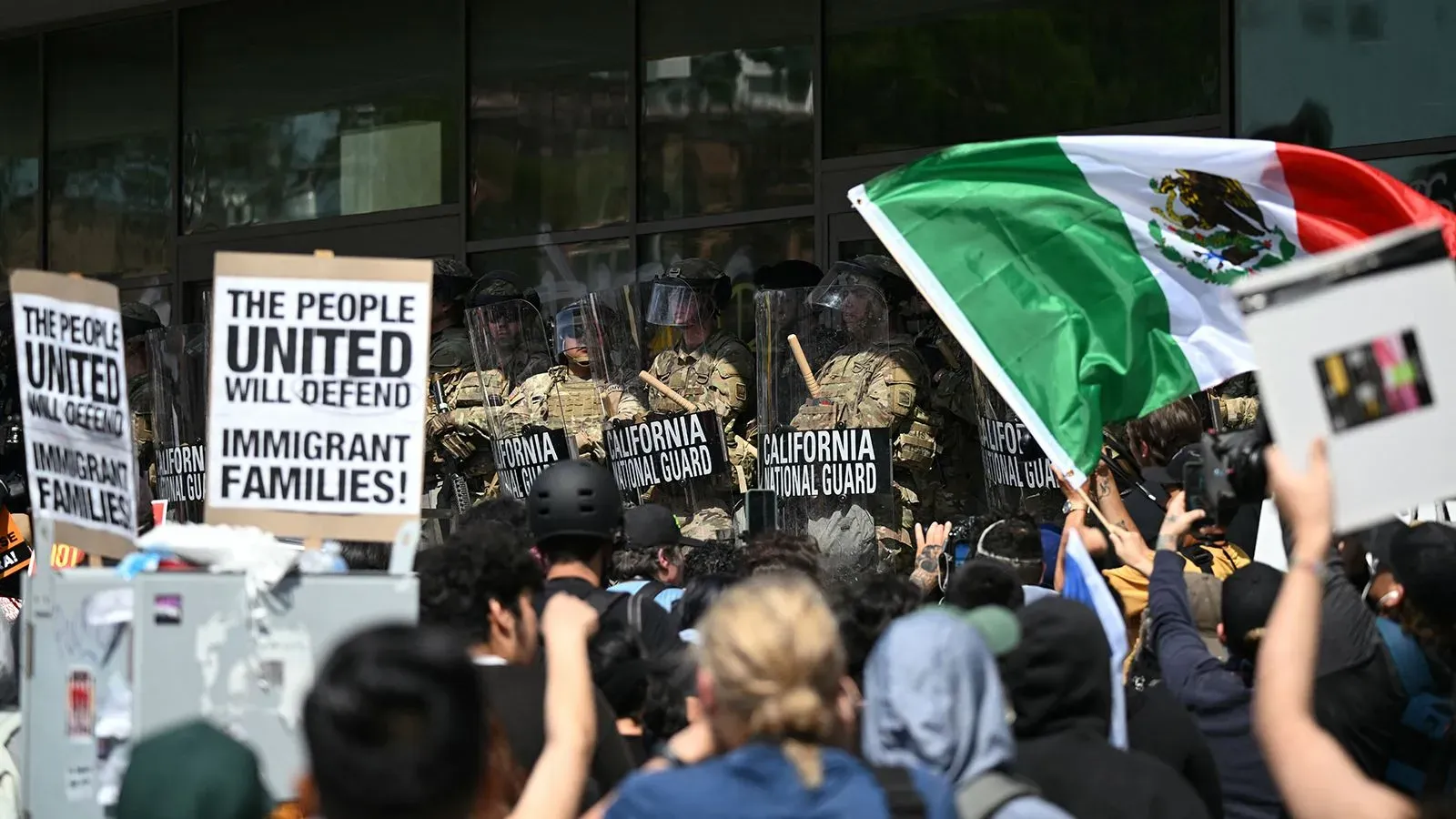 احتجاجات في لوس أنجلوس ضد إدارة الهجرة، مع وجود أفراد من الحرس الوطني يرتدون دروعًا، لافتات تدعو للدفاع عن الأسر المهاجرة.