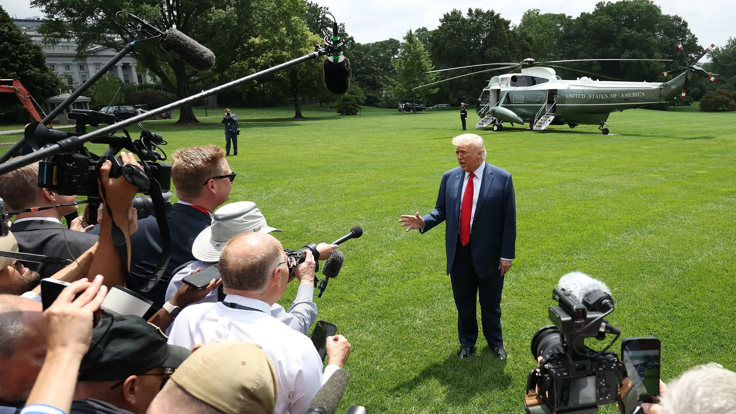 ترامب يتحدث للصحفيين في حديقة البيت الأبيض، محاطًا بكاميرات ومراسلين، مع هليكوبتر خلفه، في سياق مناقشات حول الاحتجاجات.