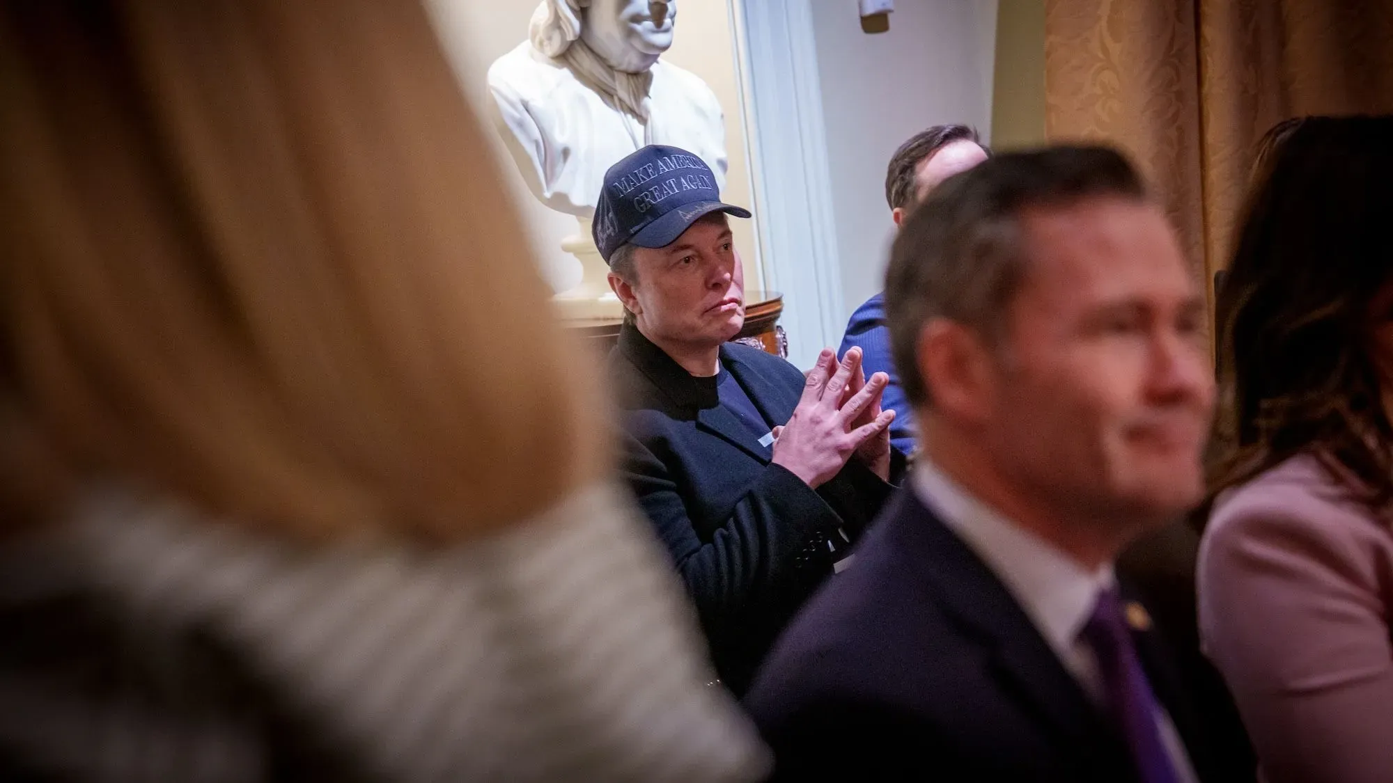 إيلون ماسك يجلس في اجتماع مجلس الوزراء، مرتديًا قبعة سوداء مكتوب عليها "اجعلوا أمريكا عظيمة مرة أخرى"، في خلفية القاعة.