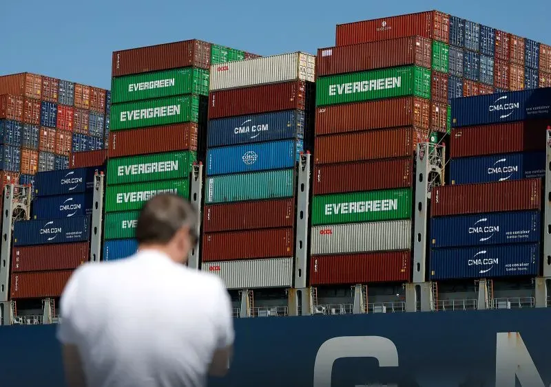 صورة لشخص يقف أمام حاويات شحن ملونة في ميناء، تعكس تأثيرات الحرب التجارية على الاقتصاد الأمريكي.