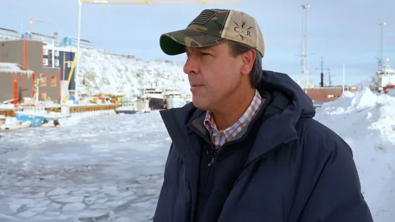 رجل يرتدي سترة ثقيلة وقبعة في بيئة ثلجية بالقرب من الميناء في نوك، غرينلاند، مع خلفية لمبانٍ ومراكب.