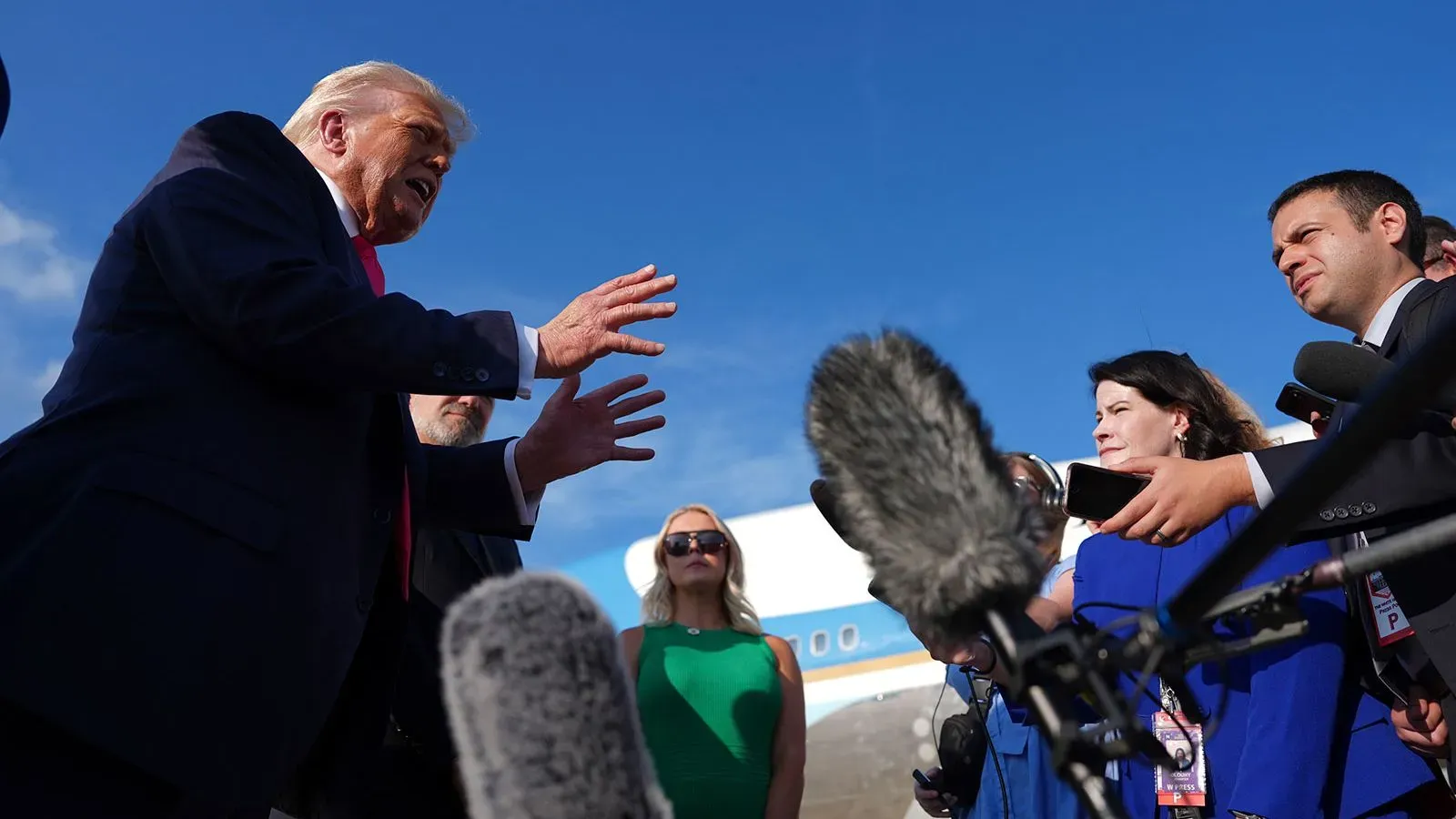 تظهر الصورة دونالد ترامب وهو يتحدث بحماس مع الصحفيين، محاطًا بمجموعة من الأشخاص، في سياق سياسي يتسم بالتوتر.