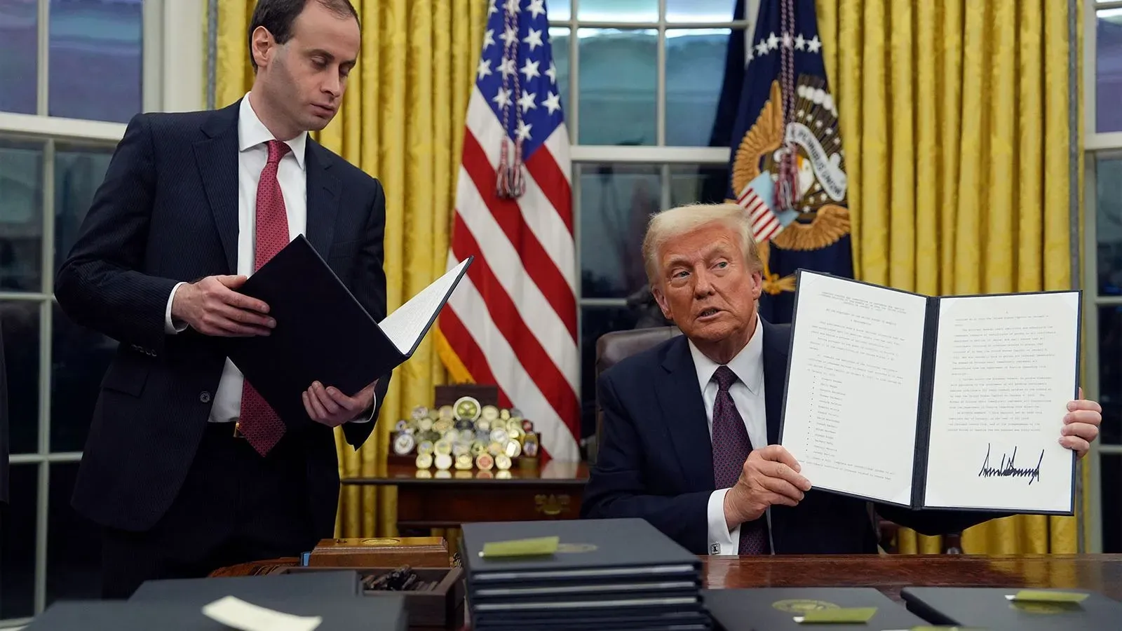 ترامب يظهر في المكتب البيضاوي وهو يحمل وثيقة موقعة، بينما ينظر مساعده إليها. الخلفية تحتوي على ستائر صفراء وشعار الرئاسة.