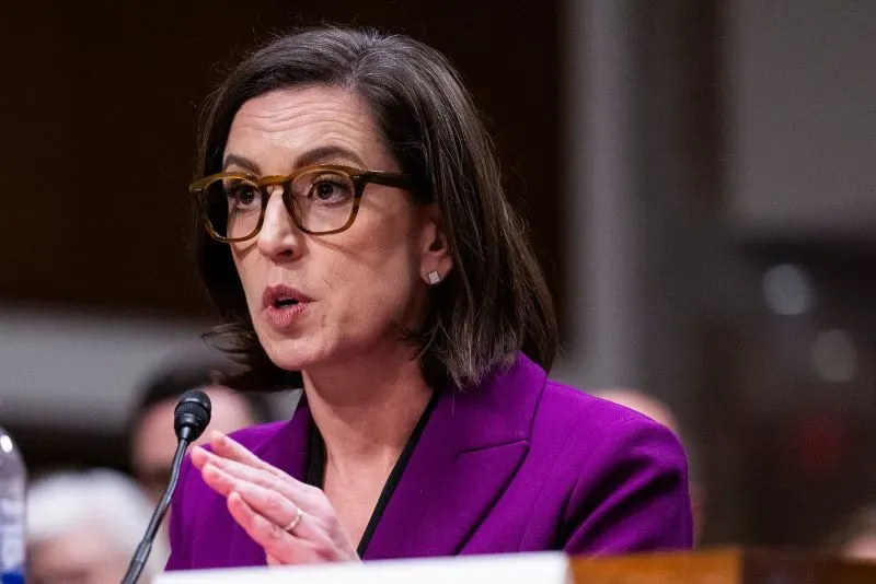 امرأة تتحدث في جلسة استماع، مرتدية سترة بنفسجية ونظارات، تعبر عن القضايا المتعلقة بفصل الموظفين الفيدراليين في إدارة ترامب.