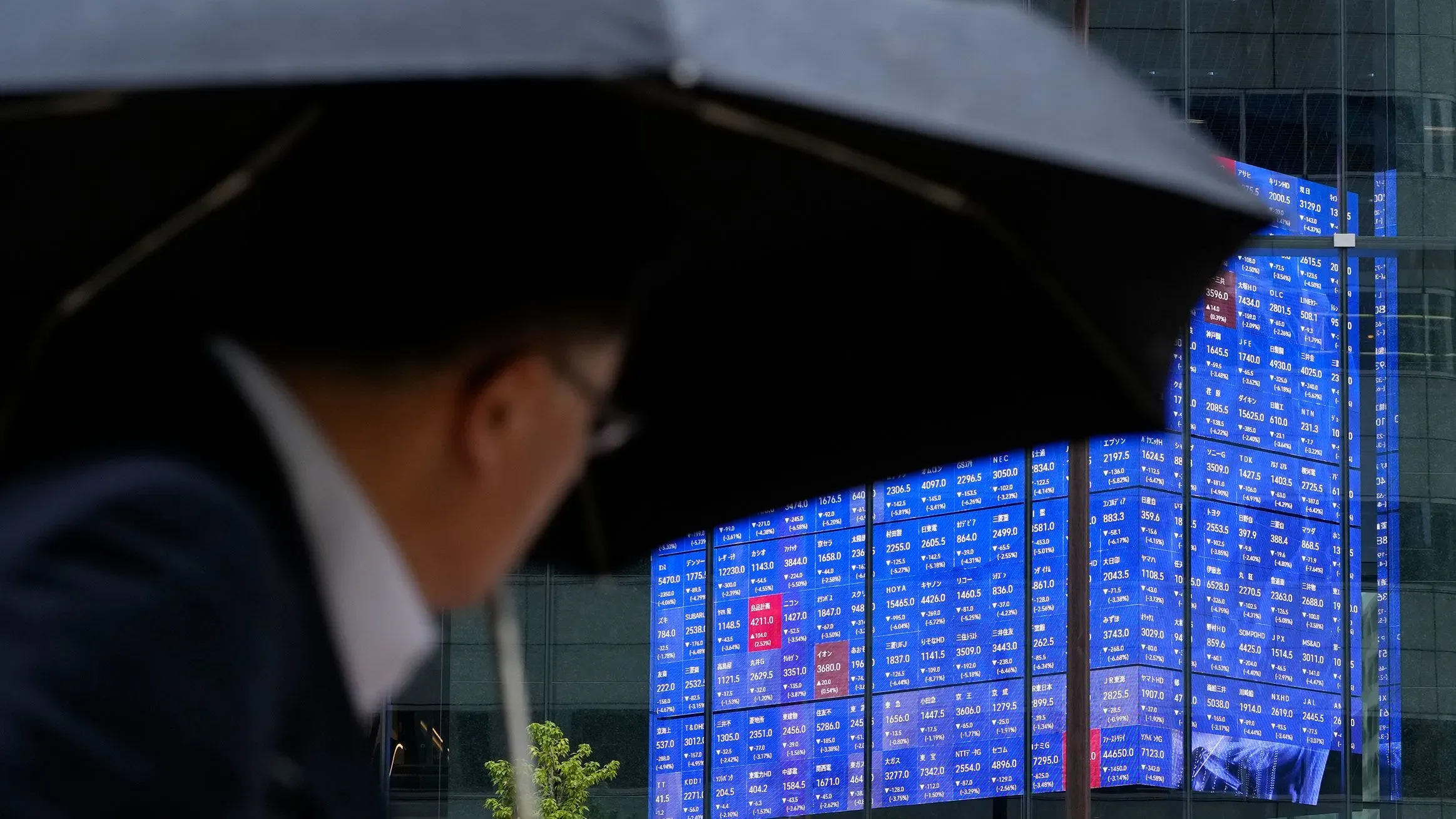 شخص يحمل مظلة أمام شاشة تعرض بيانات سوق الأسهم، مع تراجع الأسواق الآسيوية بعد فرض ترامب رسومًا جمركية جديدة.