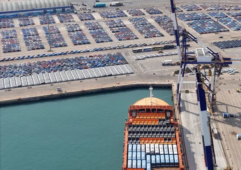 صورة لميناء يضم سفينة شحن محملة بالسيارات، مع صفوف من السيارات في الخلفية، تعكس تأثير الرسوم الجمركية على التجارة الآسيوية.