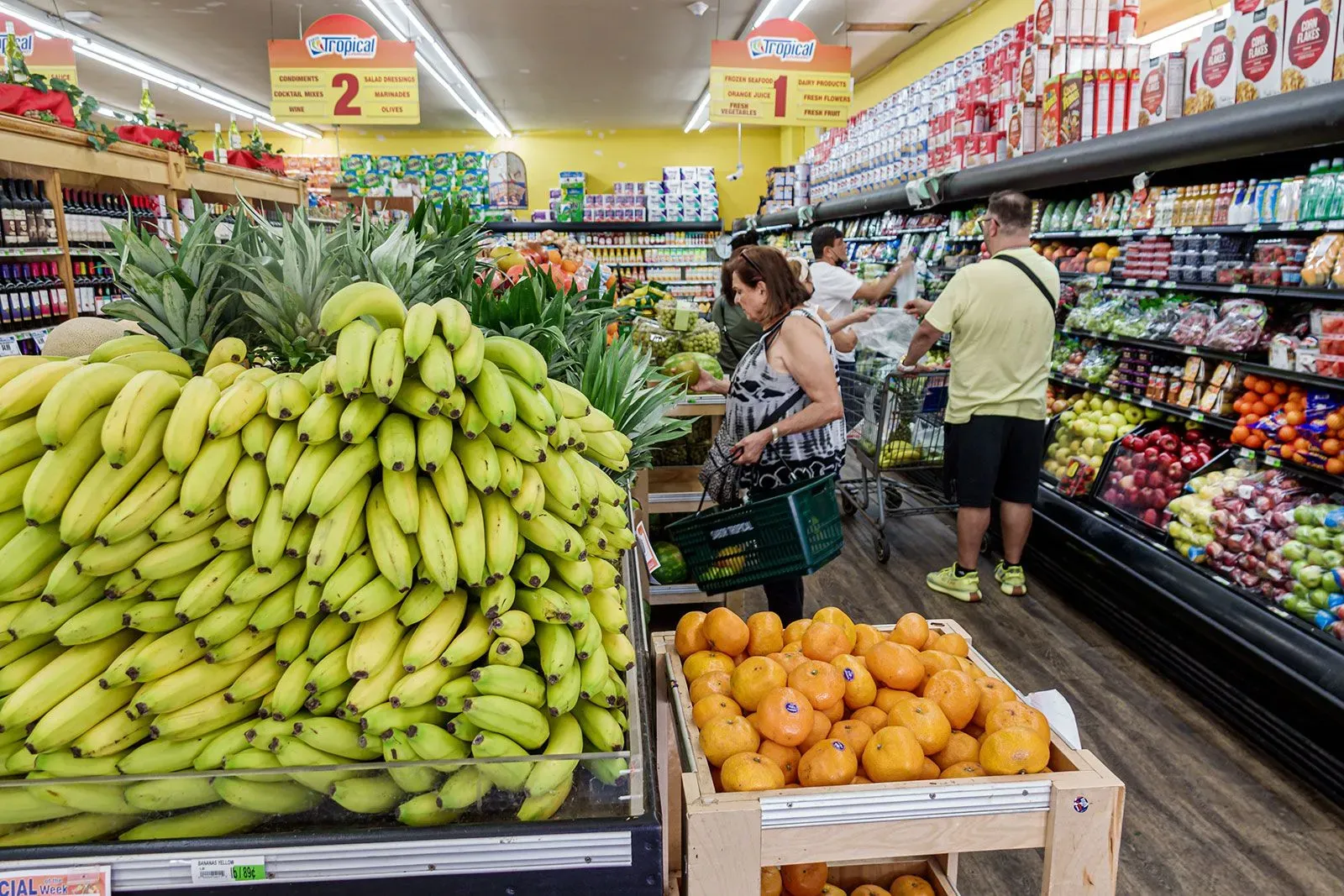 سوق بقالة مزدحم يعرض الموز والبرتقال، مع متسوقين يتجولون بين الأرفف المليئة بالفواكه والخضروات الطازجة.