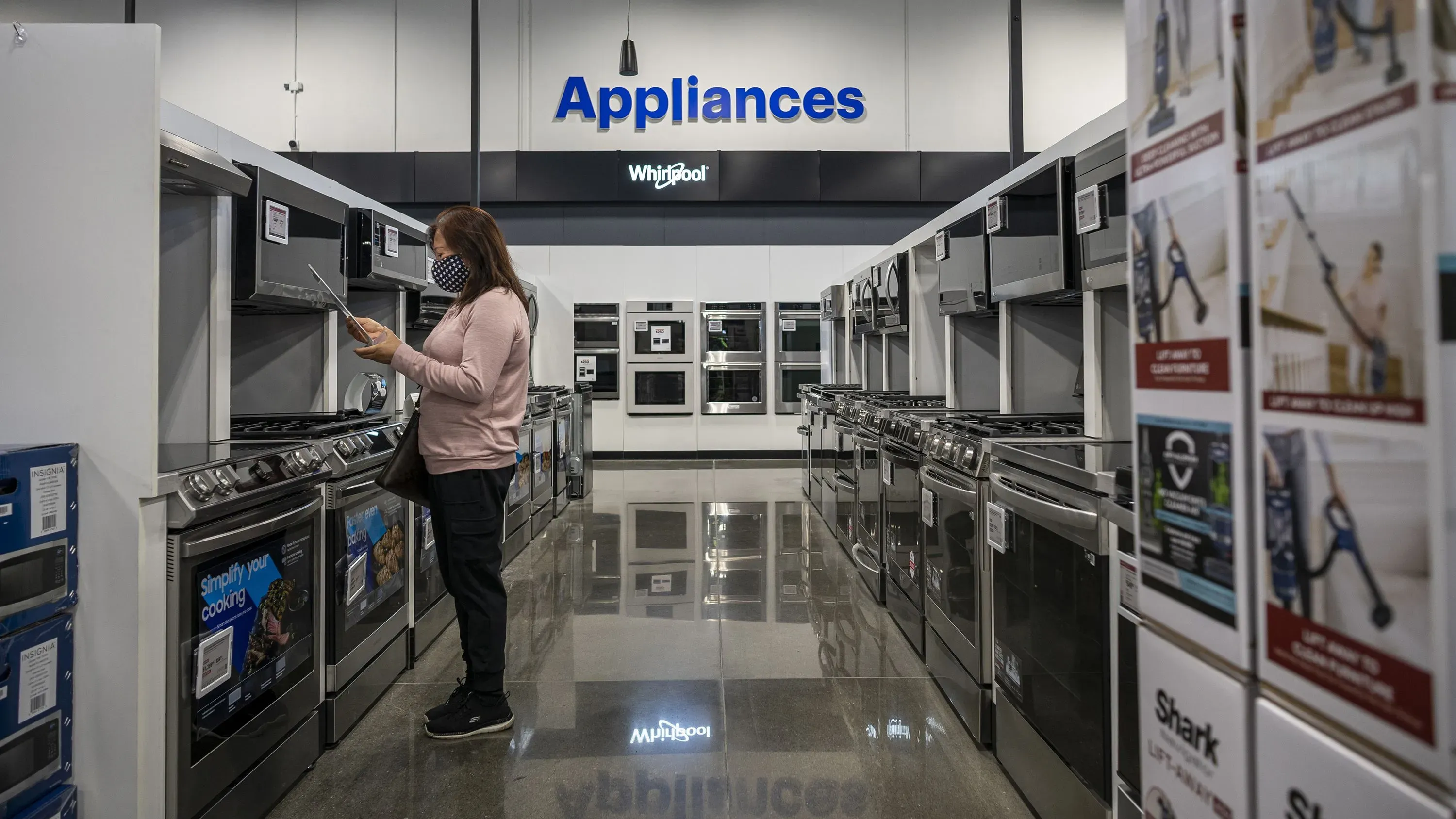 امرأة ترتدي كمامة تتفقد الأجهزة المنزلية في متجر، مع التركيز على مجموعة من الأفران والمواقد، في سياق زيادة التعريفات الجمركية على الرقائق.