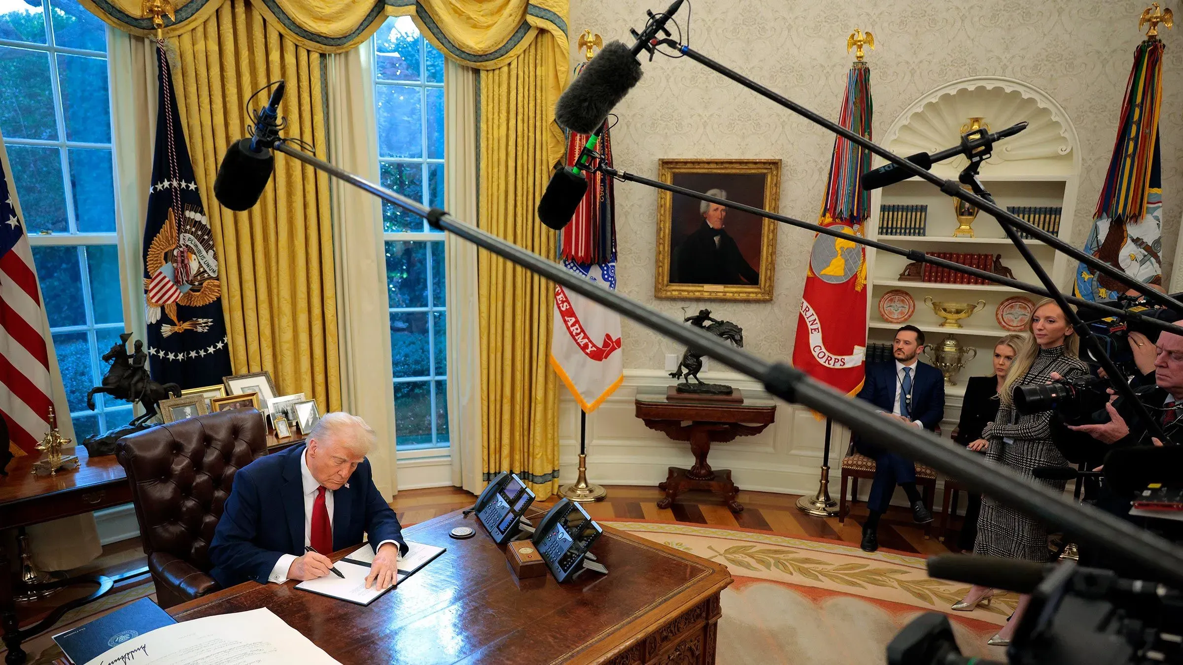 دونالد ترامب يجلس خلف مكتبه في المكتب البيضاوي، يوقع وثيقة، محاطًا بكاميرات وصحفيين، بينما يتحدث عن سياسات التنوع والمساواة.