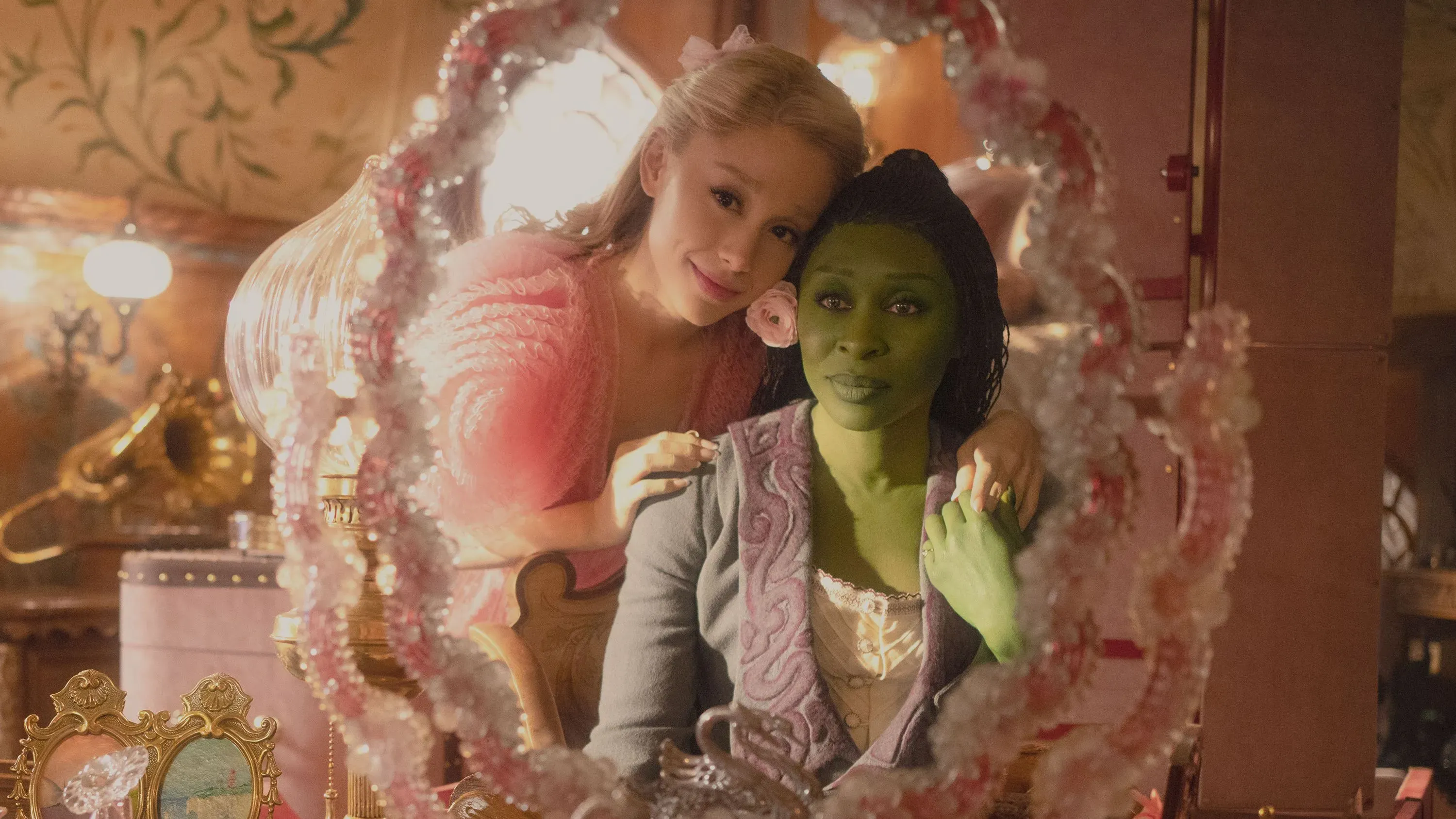 امرأتان تجلسان معًا أمام مرآة، واحدة بشعر أشقر ترتدي ملابس وردية، والأخرى بشرة خضراء ترتدي ملابس تاريخية، تعكسان روح التعاون في صناعة السينما.