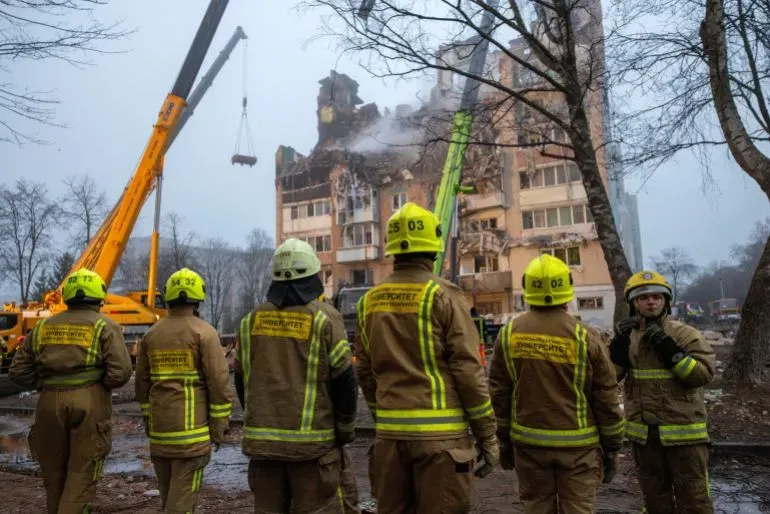 رجال إطفاء يرتدون زي العمل يقفون أمام مبنى مدمر، حيث تُستخدم رافعة لإزالة الأنقاض في موقع الهجوم الروسي على أوكرانيا.