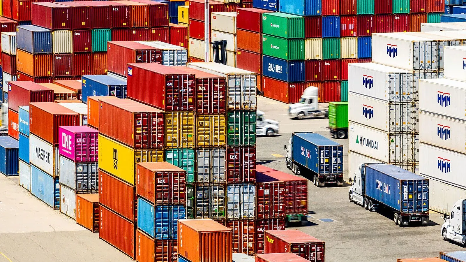 حاويات شحن ملونة مكدسة في ميناء، مع شاحنات تسليم تتحرك بينها، تعكس تأثير التعريفات الجمركية الجديدة على التجارة العالمية.