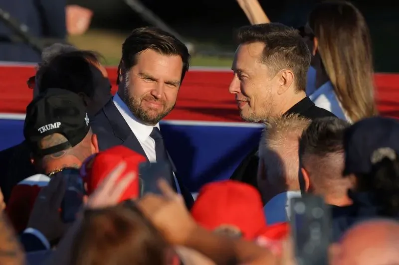 إيلون ماسك يتحدث مع شخص آخر في تجمع سياسي، مع وجود حشود من المؤيدين في الخلفية، مما يعكس تأثيره المتزايد في السياسة الأمريكية.