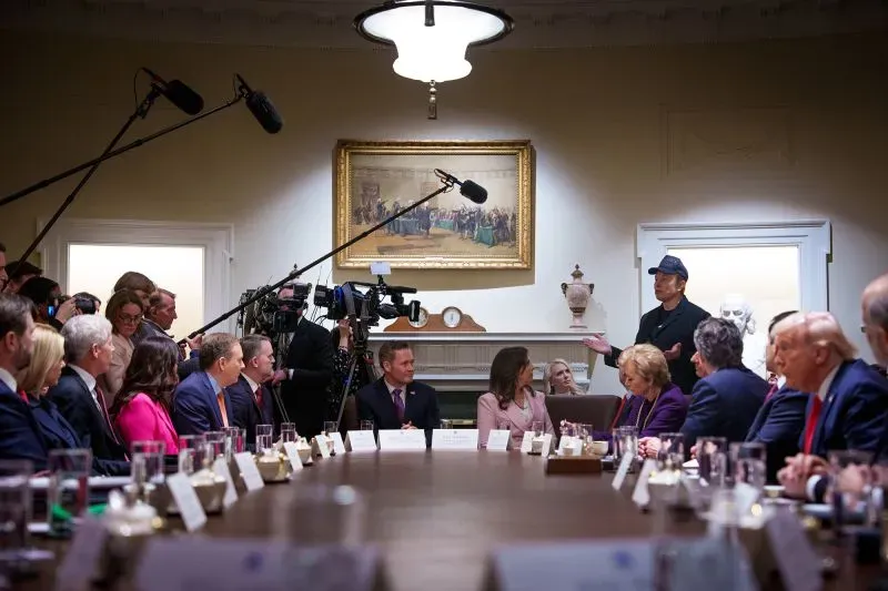 اجتماع في البيت الأبيض يضم شخصيات سياسية وإعلامية، مع وجود كاميرات وتصوير، حيث يتحدث إيلون ماسك في المقدمة.