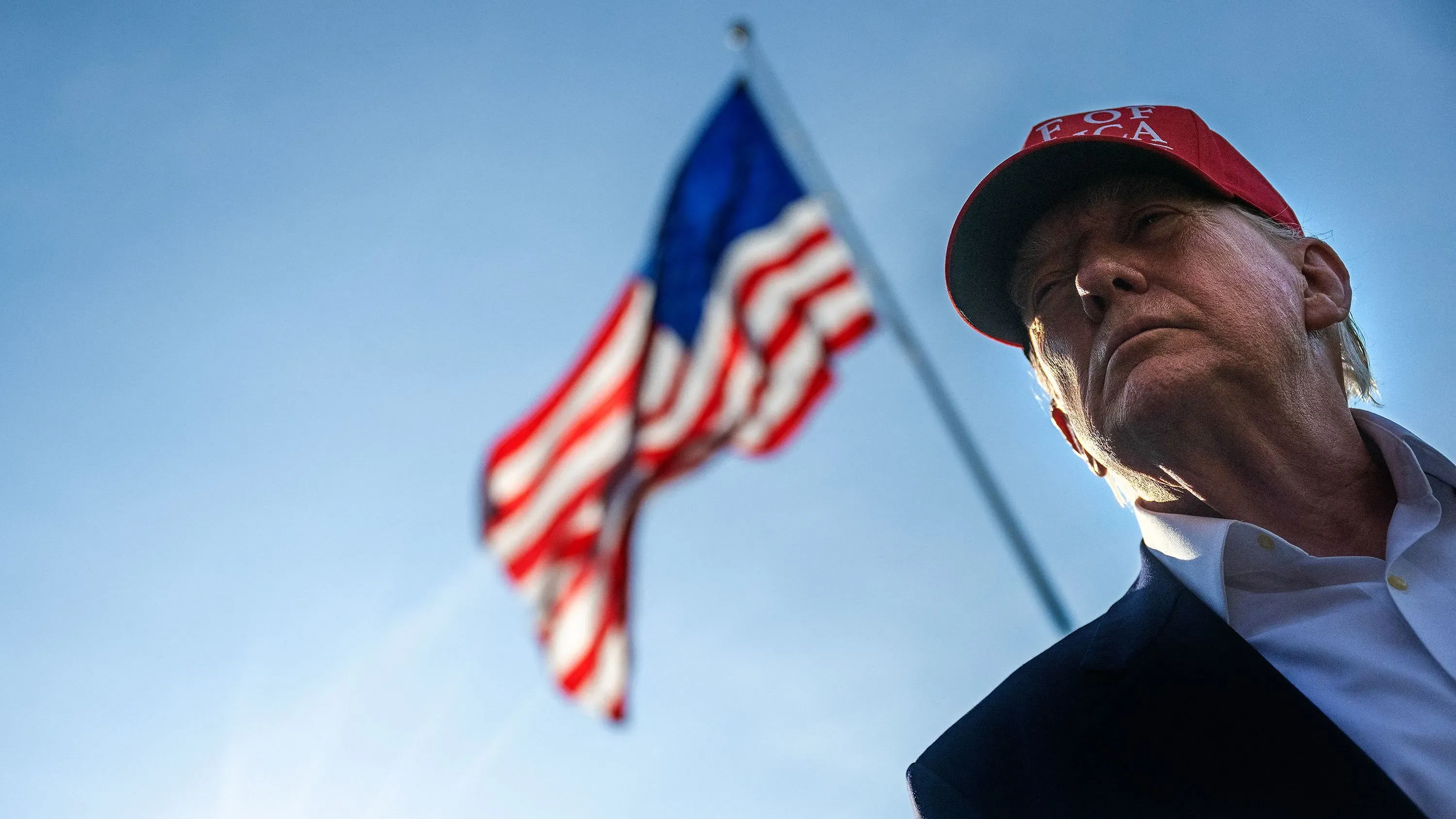 صورة لدونالد ترامب يرتدي قبعة حمراء تحمل عبارة "Make America Great Again"، يقف أمام علم الولايات المتحدة في سماء زرقاء.