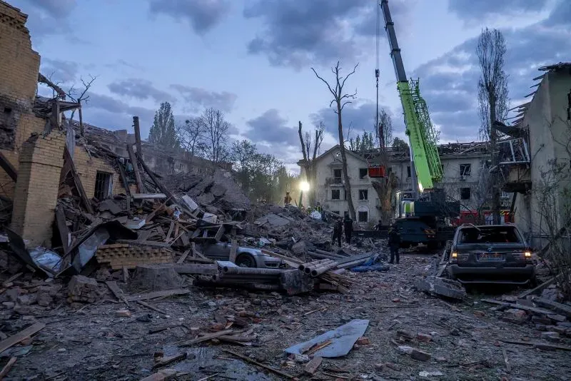 تظهر الصورة مشهدًا مدمرًا في منطقة حضرية، حيث تهدمت المباني بفعل القصف، مع وجود عمال إنقاذ ومركبات في الموقع.