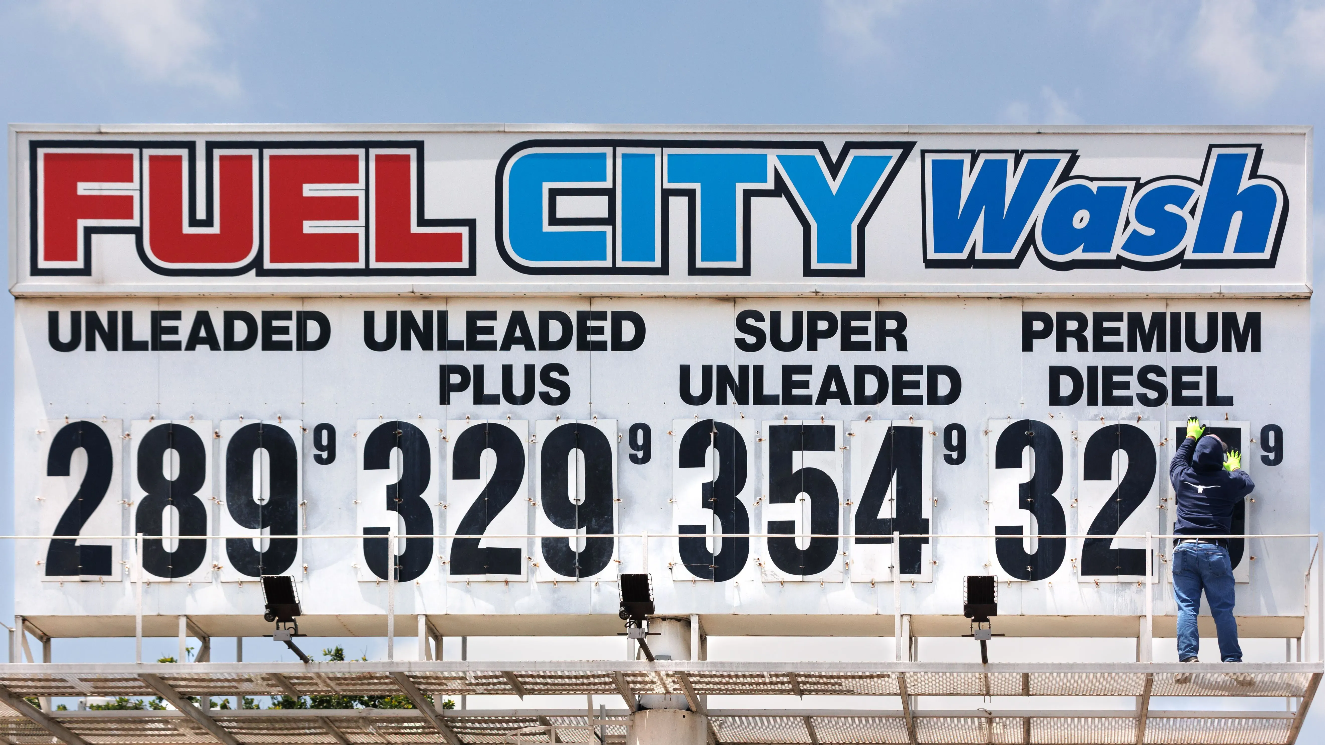 لوحة إعلانات لمحطة وقود تعرض أسعار البنزين بأنواعه المختلفة، مع شخص يقوم بتحديث الأسعار. تعكس الصورة تأثير أسعار النفط على السوق.