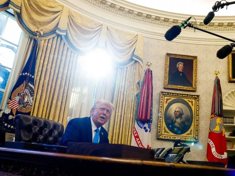 دونالد ترامب يتحدث في المكتب البيضاوي، محاطًا بالستائر الذهبية والرموز الرئاسية، تحت أضواء الكاميرات.