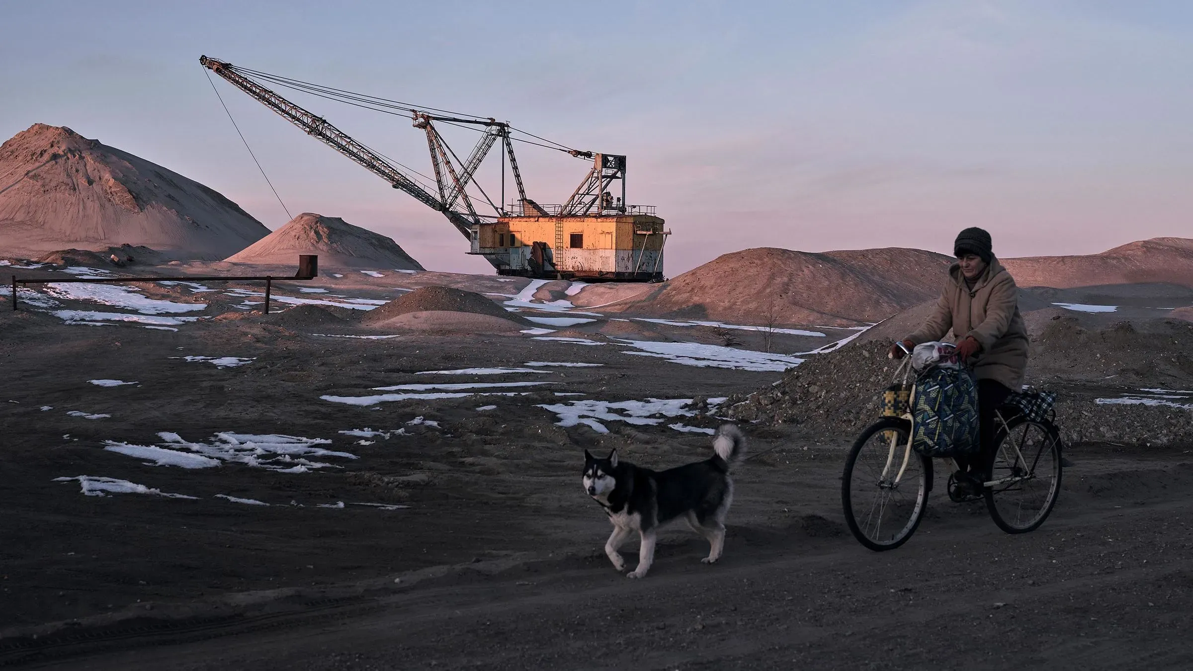 امرأة تركب دراجة قرب موقع تعدين، مع وجود كلب يرافقها، وآلة حفر كبيرة في الخلفية، تعكس النشاط الصناعي في المنطقة.
