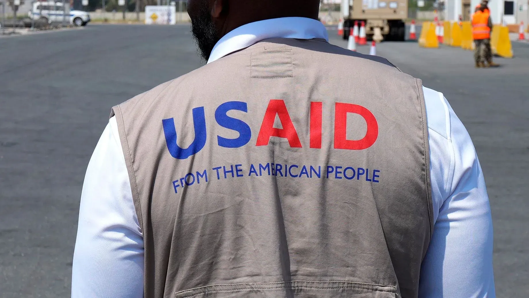 موظف من الوكالة الأمريكية للتنمية الدولية (USAID) يرتدي سترة تحمل شعار الوكالة، في موقع عمل خارجي، مع خلفية تظهر منطقة مفتوحة.