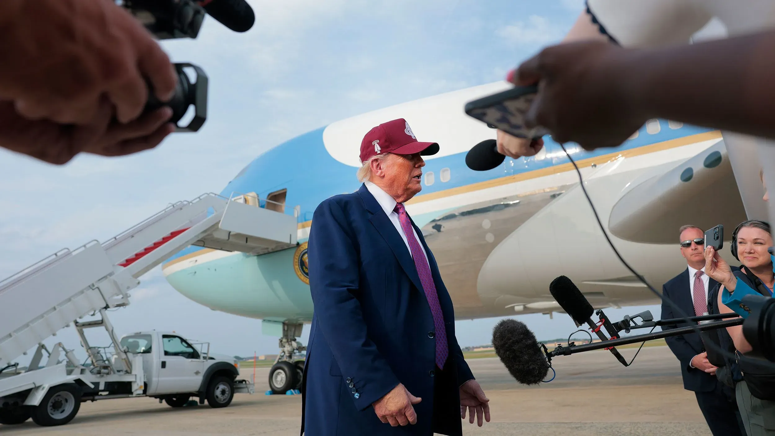 الرئيس ترامب يتحدث للصحفيين أمام طائرته، مع وجود معدات إعلامية ومراسلين يلتقطون الصور في خلفية مشهد يعكس التوترات السياسية.