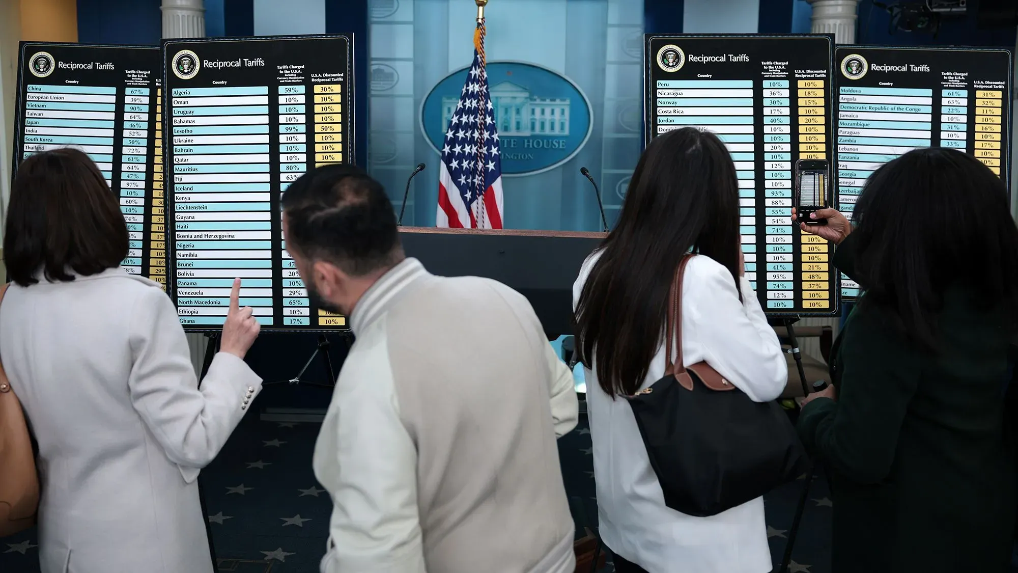 شخصان يتفقدان لوحات تعرض معدلات الرسوم الجمركية المتبادلة في مؤتمر صحفي بالبيت الأبيض، مع العلم الأمريكي خلفهم.