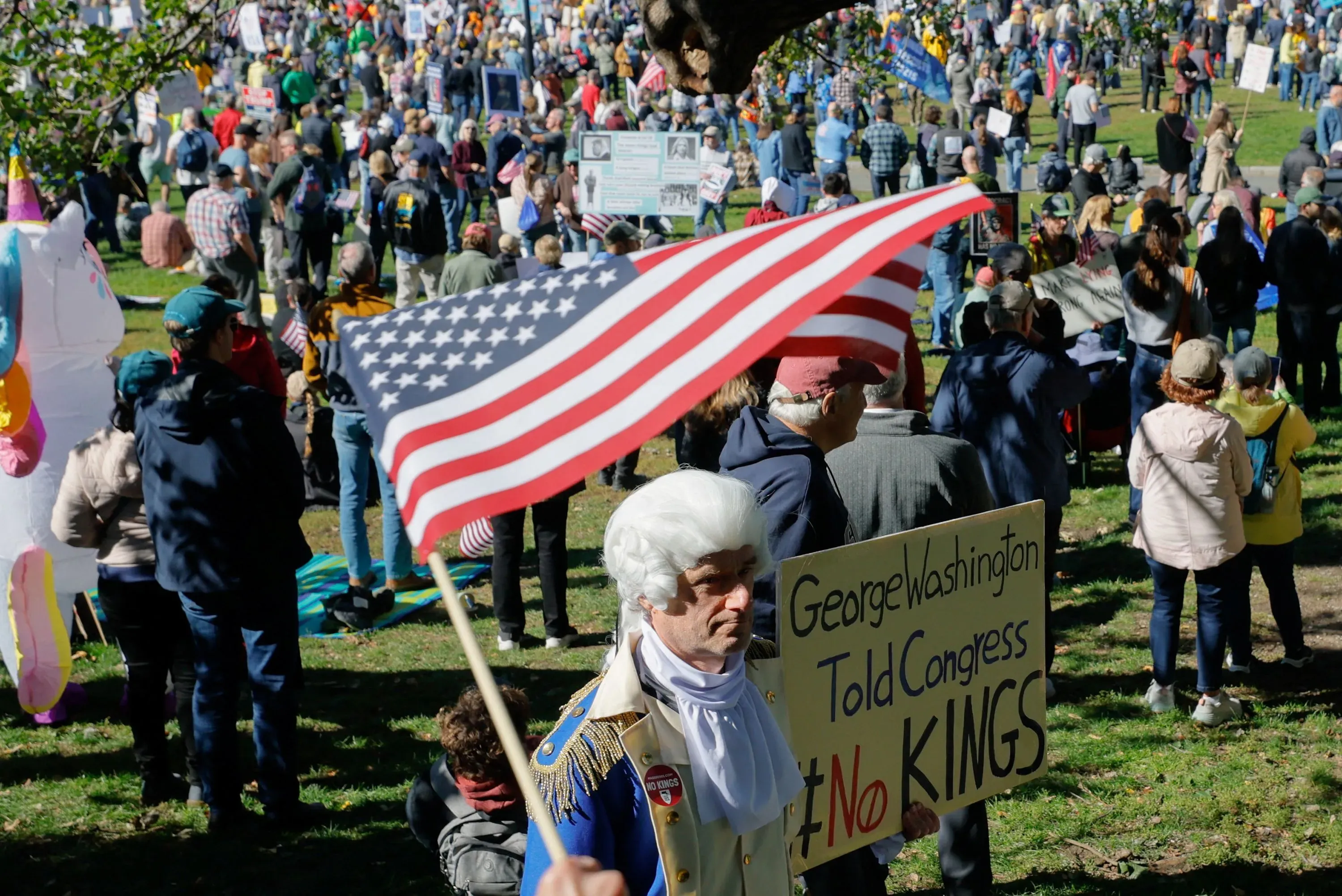 محتجون يحملون لافتات وأعلامًا أمريكية في مسيرة ضد الحكم الملكي، مع شخصية ترتدي زي جورج واشنطن.