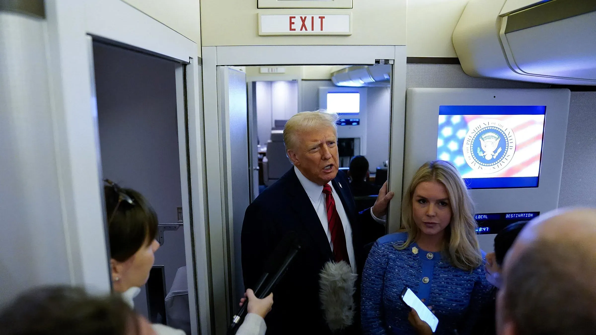 دونالد ترامب يتحدث مع الصحفيين في ممر الطائرة الرئاسية، مع وجود شاشة تعرض شعار الحكومة الأمريكية خلفه.
