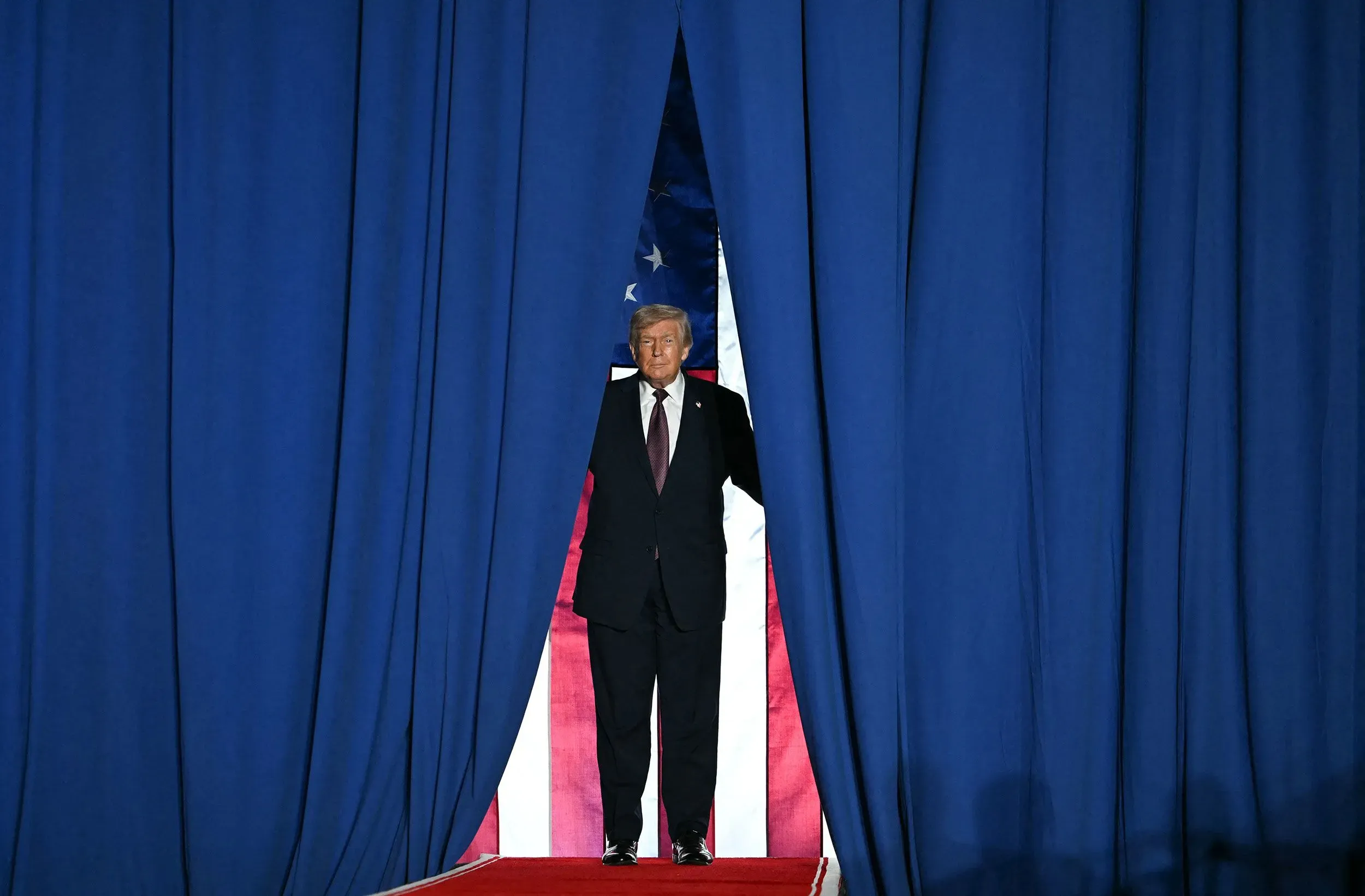ترامب يقف أمام ستارة زرقاء، مع العلم الأمريكي خلفه، استعدادًا للظهور في حدث سياسي. تعكس الصورة نشاطه في جمع التبرعات.