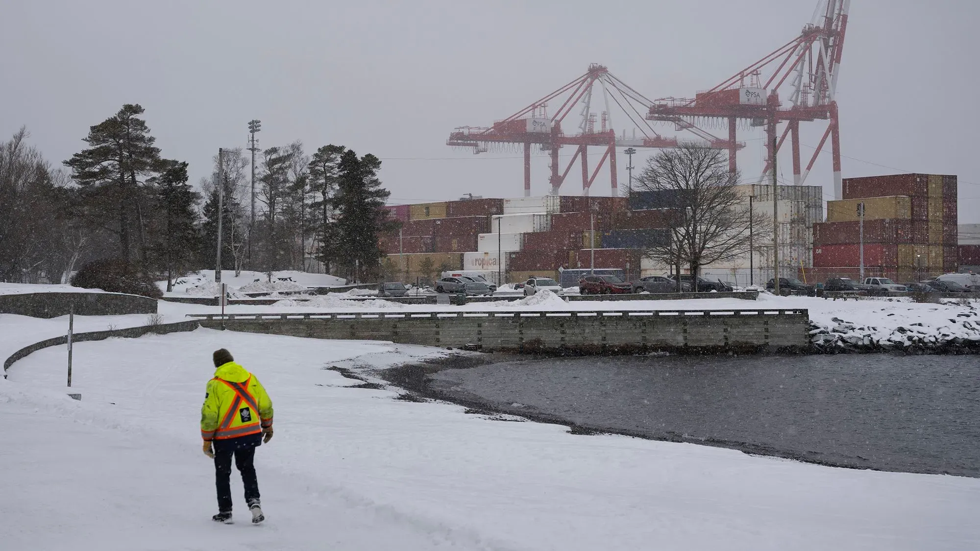عامل يرتدي سترة عاكسة يمشي على شاطئ مغطى بالثلوج، خلفه حاويات وشاحنات في ميناء أثناء تساقط الثلوج.