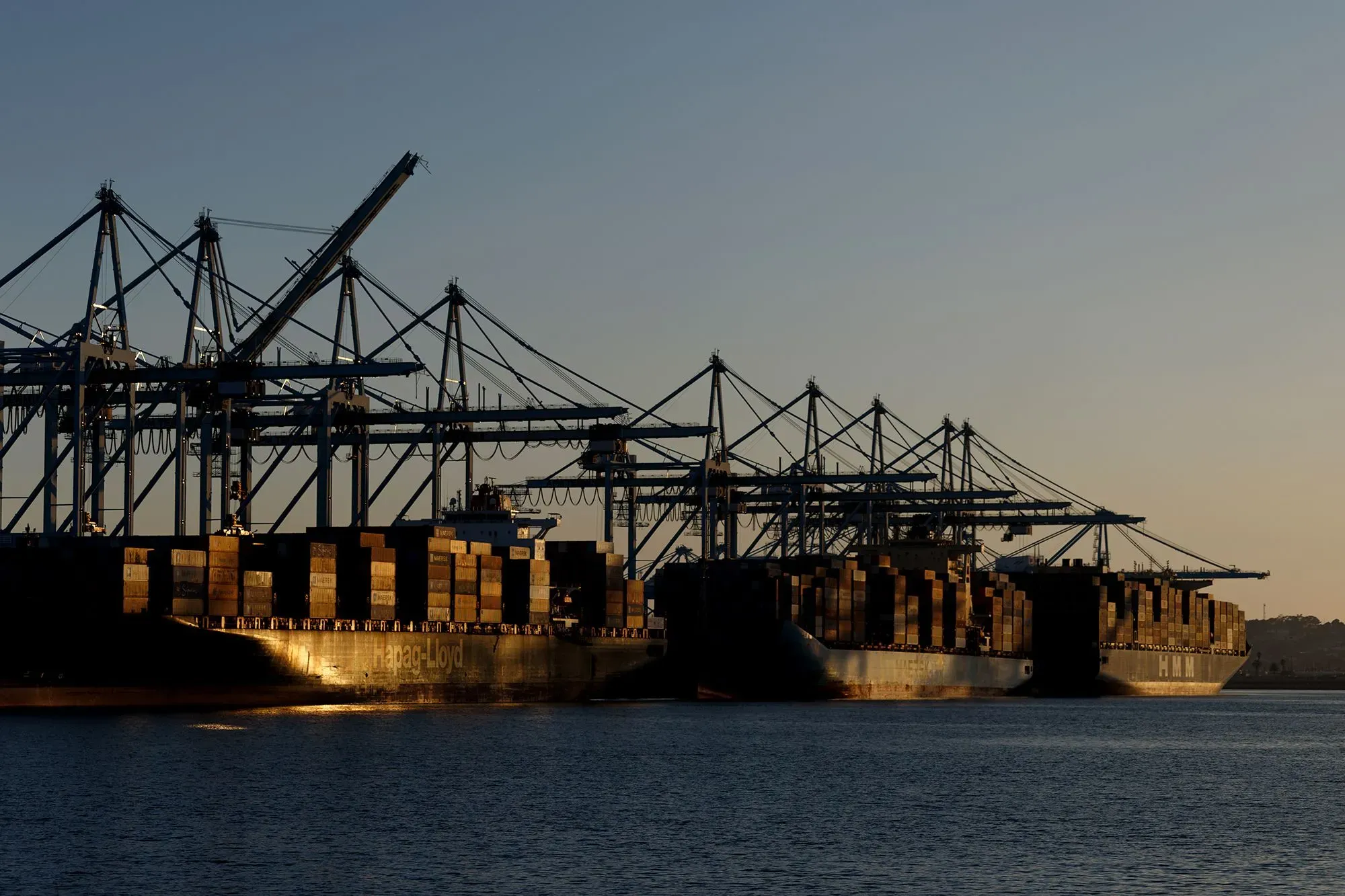 سفن شحن محملة بالحاويات في ميناء، مع معدات تحميل بارزة، تعكس تأثير الرسوم الجمركية على التجارة خلال الإغلاق الحكومي.