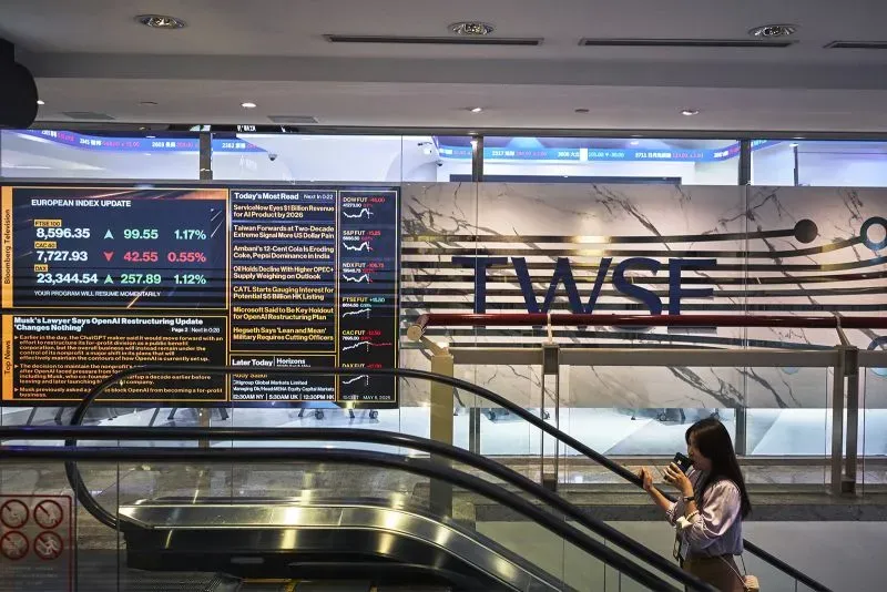 امرأة تستخدم هاتفها في ممر سوق الأسهم التايواني، مع شاشة تعرض مؤشرات السوق وأرقام التداول، تشير إلى تقلبات العملات.