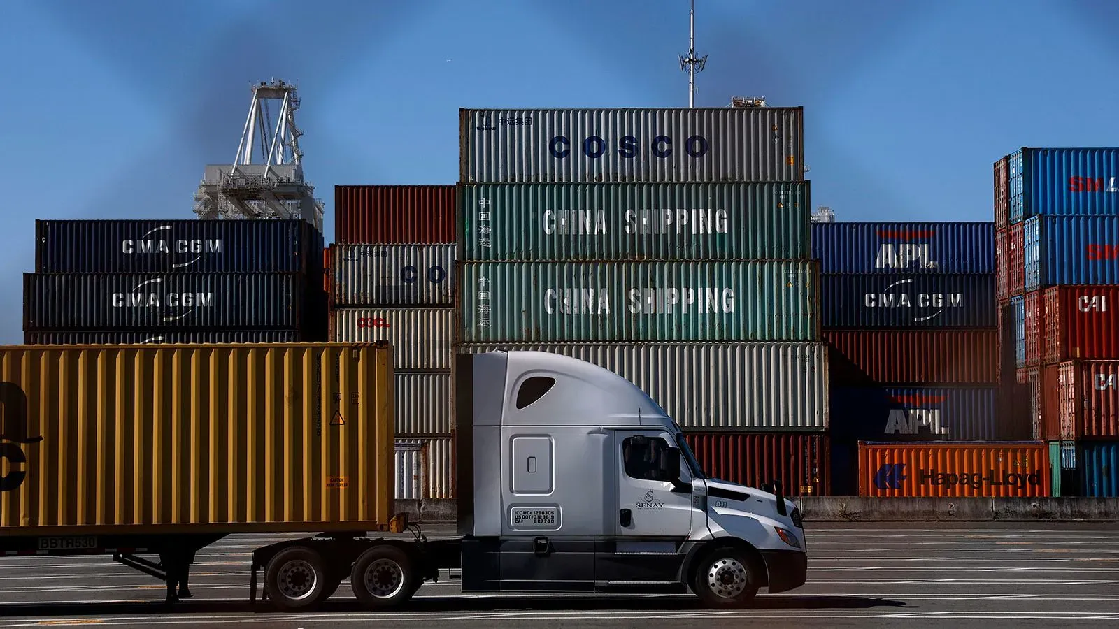 شاحنة نقل أمام حاويات شحن متعددة الألوان في ميناء، تعكس تأثير التعريفات الجمركية على الواردات الأمريكية.