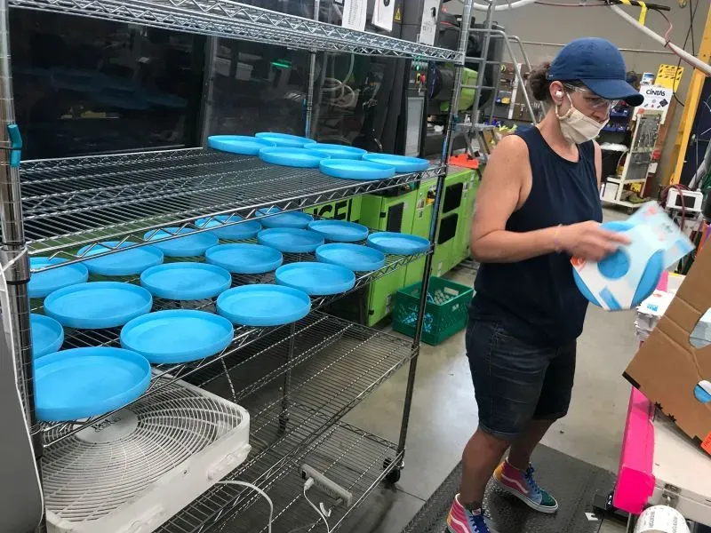 امرأة ترتدي قناعًا تعمل في مصنع، حيث تقوم بتعبئة ألعاب الحيوانات الأليفة، مع وجود أطباق زرقاء مرتبة على رفوف خلفها.