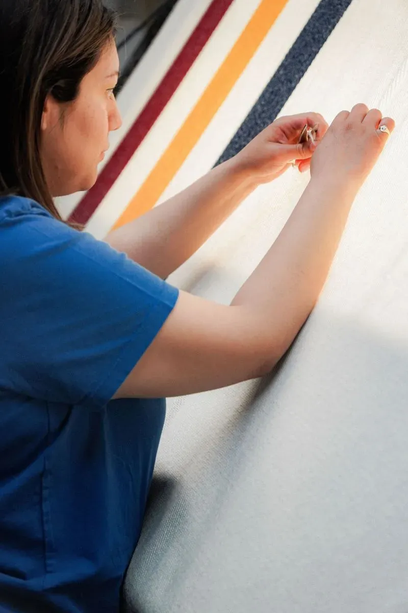 امرأة تعمل على خياطة بطانية في مطحنة فاريبولت، مع التركيز على تفاصيل الألوان والنمط، مما يعكس الحرفية التقليدية في صناعة البطانيات.